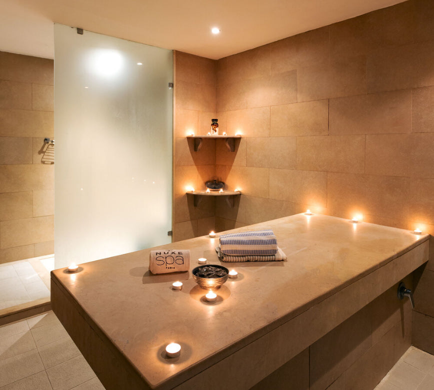 Salle de massage élégante du spa NUXE à l’Hôtel Les Berges du Lac – Concorde, composée d’une large table de soin minérale entourée de bougies, d’étagères décoratives et d’un espace douche vitré, baignée d’une lumière douce pour une ambiance chaleureuse, sereine et propice à la détente.
