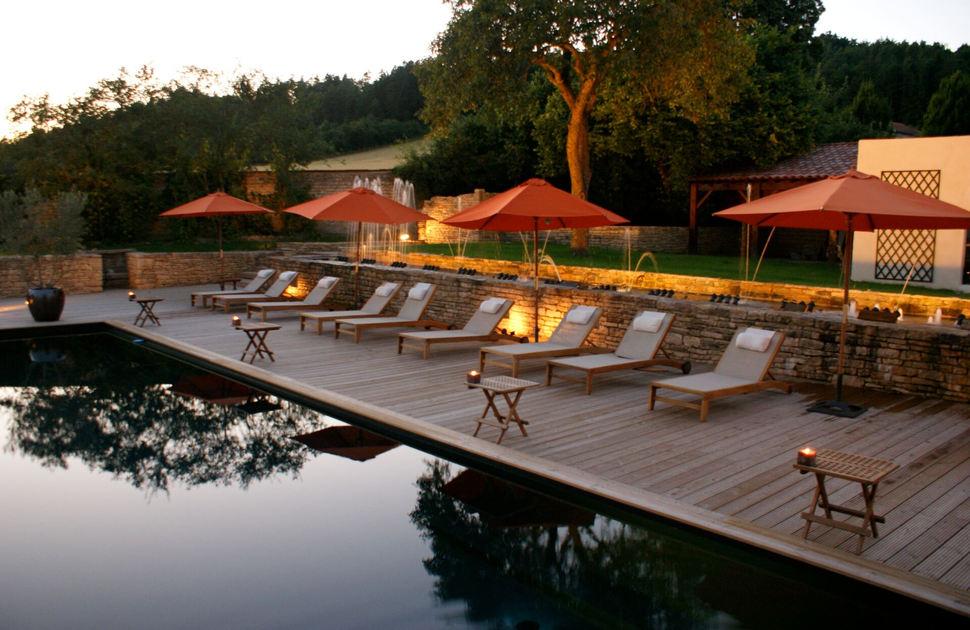 Un espace extérieur chaleureux et raffiné à la tombée du jour, où une piscine élégante se fond dans un décor naturel apaisant. Entouré de transats en bois, de parasols colorés et de touches lumineuses, ce lieu invite à un moment de détente privilégié dans une atmosphère à la fois intime et conviviale.