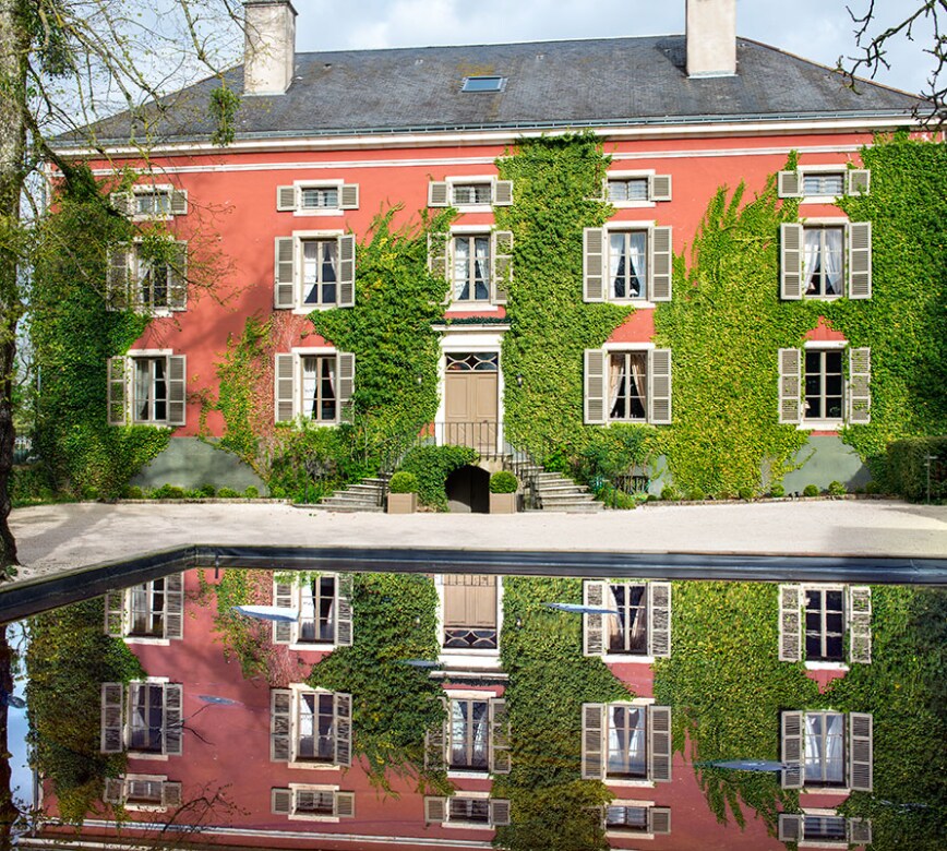  Superbe maison de charme aux murs rosés ornés de végétation grimpante, avec volets clairs et entrée symétrique, se reflétant harmonieusement dans un bassin, dans une atmosphère sereine et élégante.
