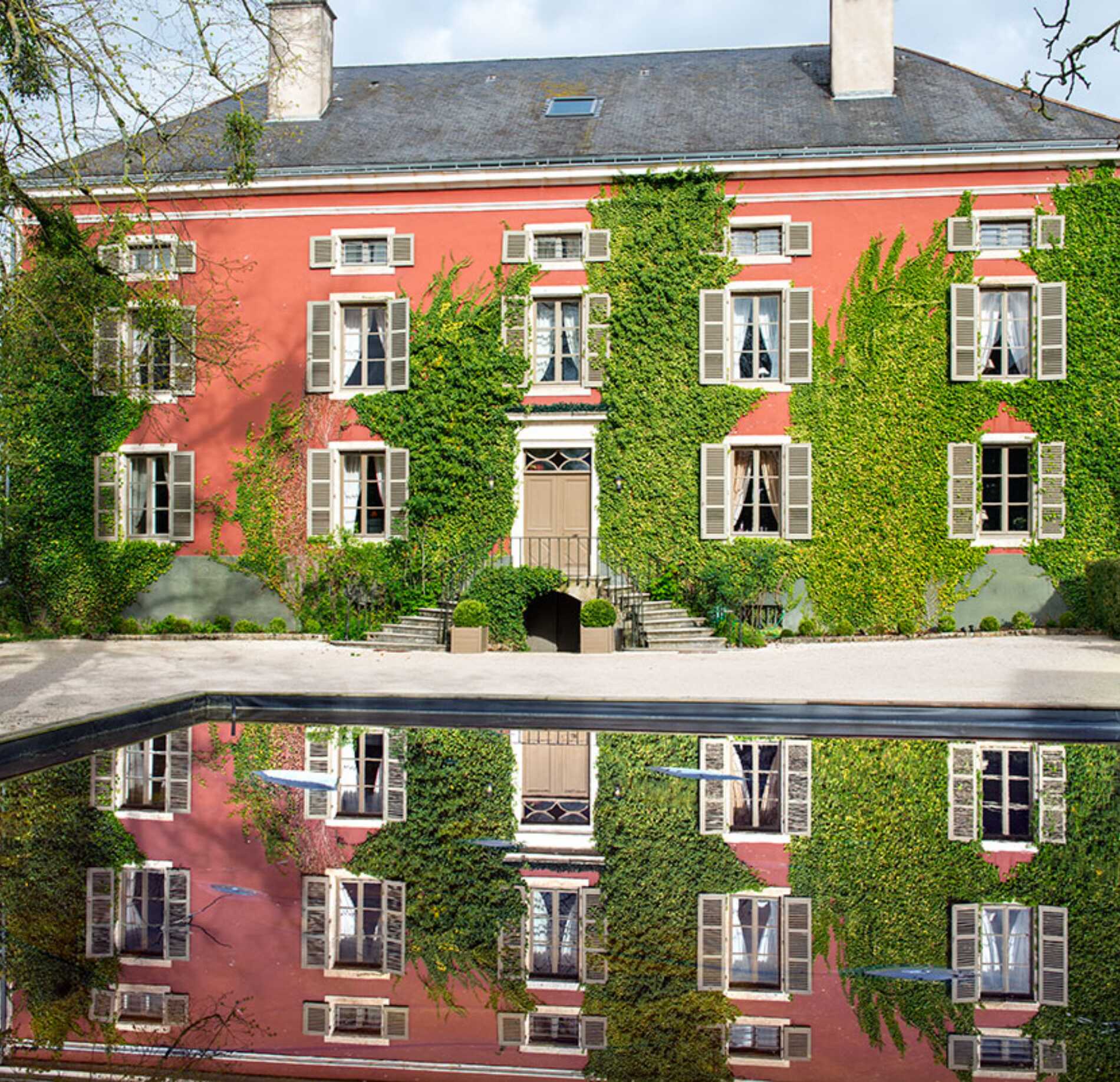 Élégante demeure de caractère aux façades rosées, élégamment habillées de lierre verdoyant, ouvrant sur un bassin calme reflétant le bâtiment, dans un cadre paisible et raffiné.