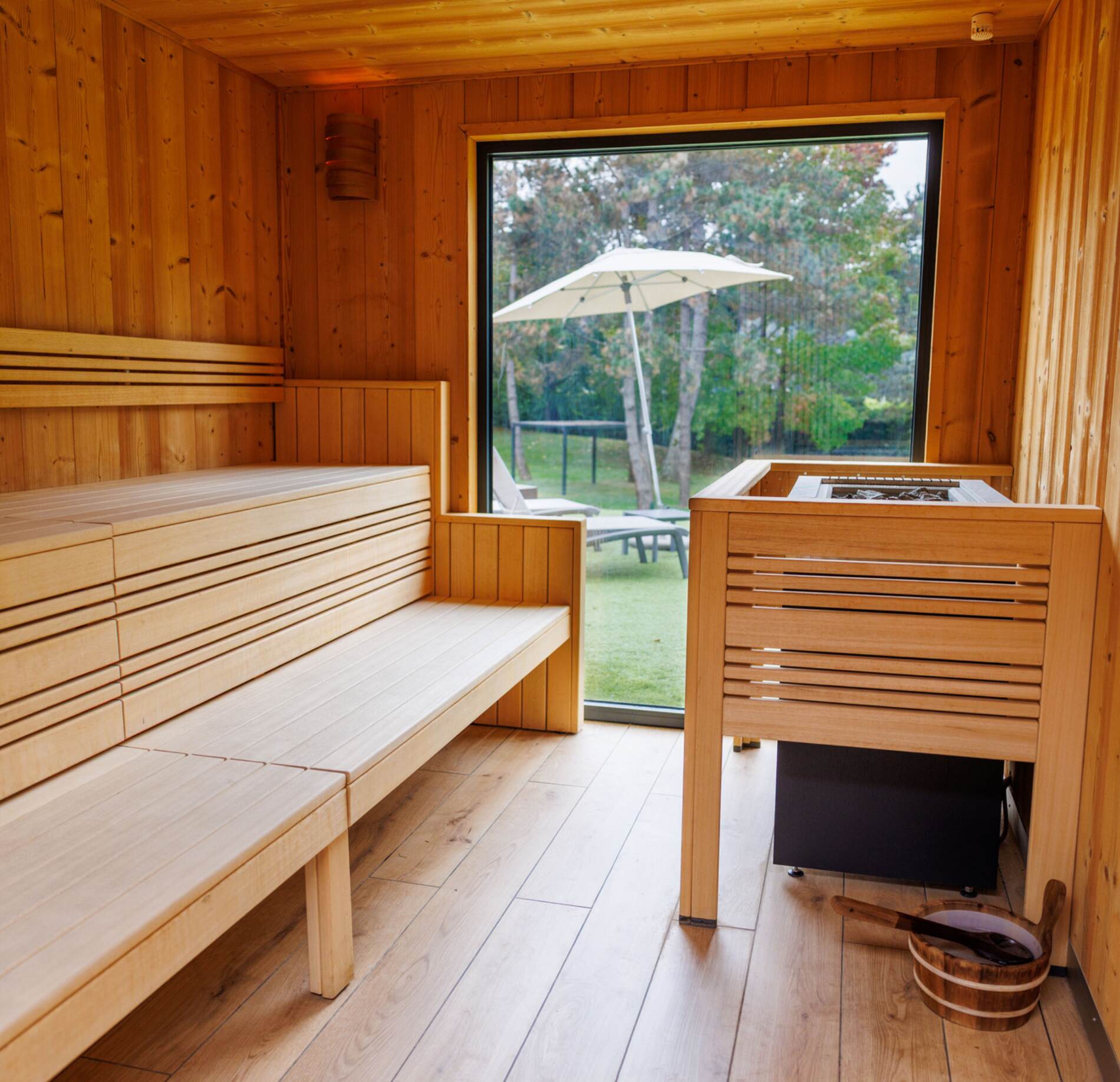 Sauna élégant entièrement habillé de bois clair, avec banquettes épurées et poêle central, ouvert sur une grande baie vitrée offrant une vue apaisante sur un jardin verdoyant, créant une atmosphère lumineuse et relaxante.