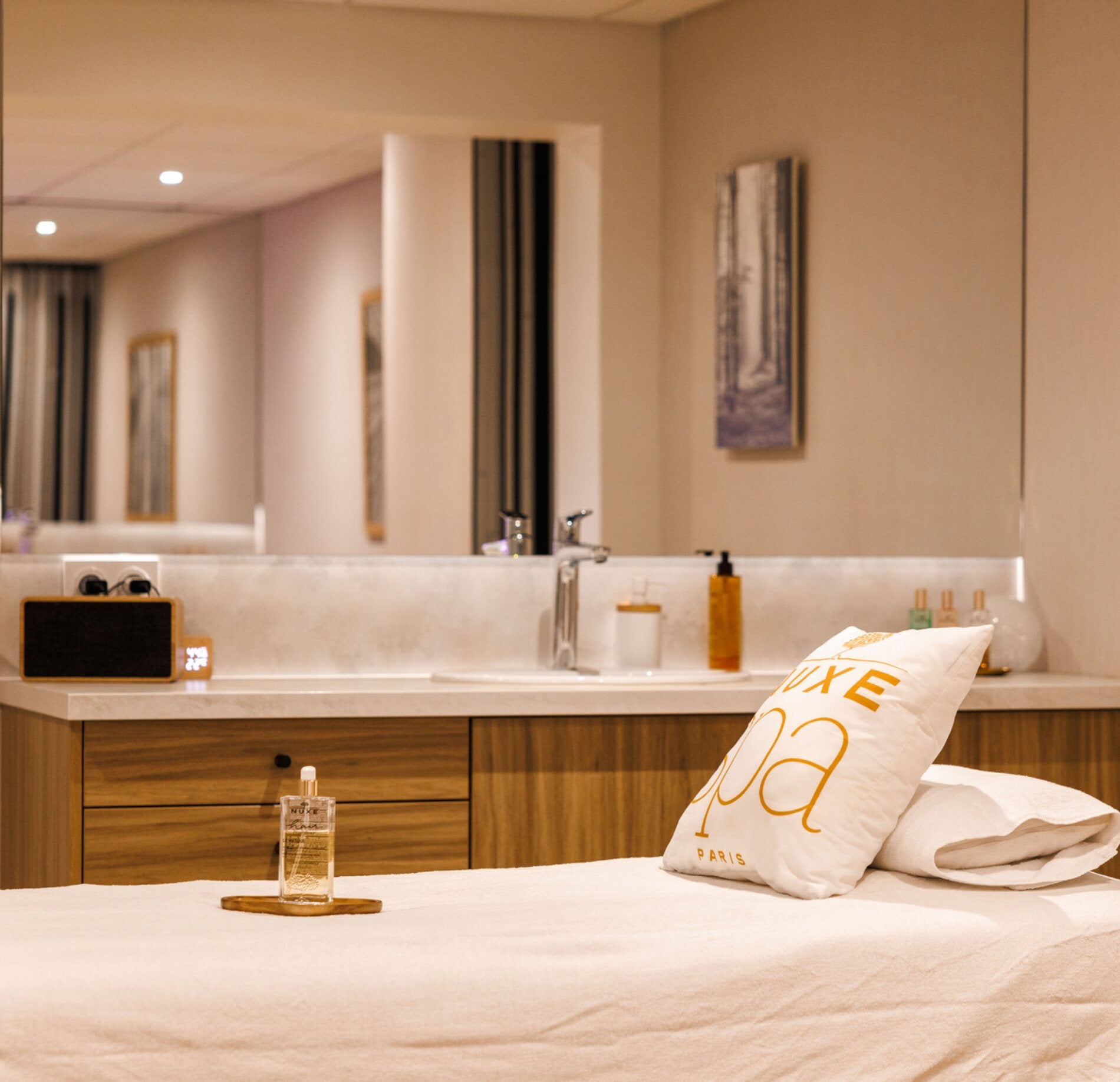 Cabine de soin élégante du spa NUXE, avec table de massage confortable, linge soigneusement disposé et coussin signature, complétée par un espace lavabo aux finitions bois et éclairage doux, créant une atmosphère apaisante et raffinée. 