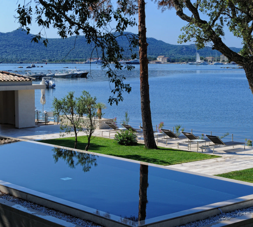 Piscine extérieure à débordement du spa NUXE à l’Hôtel Don César, bordée de transats élégants et d’un jardin verdoyant, offrant une vue panoramique sur une mer paisible avec bateaux et collines en arrière-plan, dans une atmosphère lumineuse, sereine et raffinée.