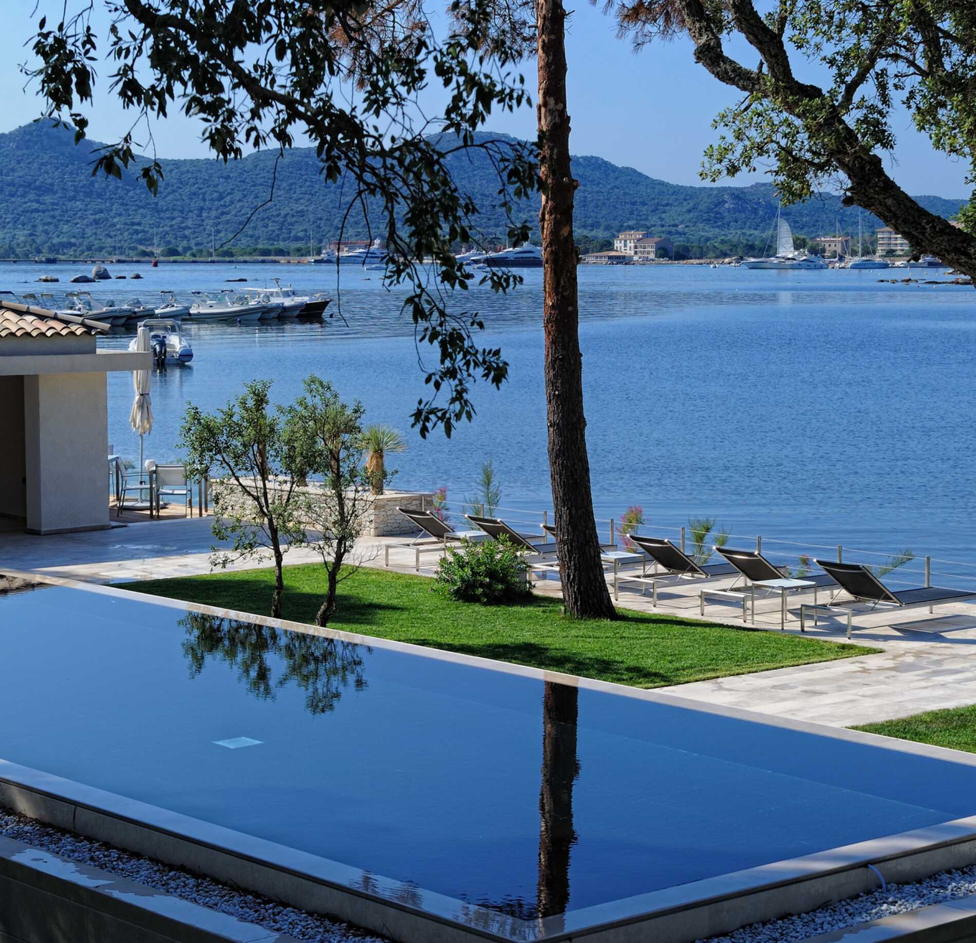 Piscine extérieure à débordement du spa NUXE à l’Hôtel Don César, bordée de transats élégants et d’un jardin soigné, offrant une vue spectaculaire sur une mer calme avec bateaux et collines en arrière-plan, dans une atmosphère lumineuse et raffinée. 