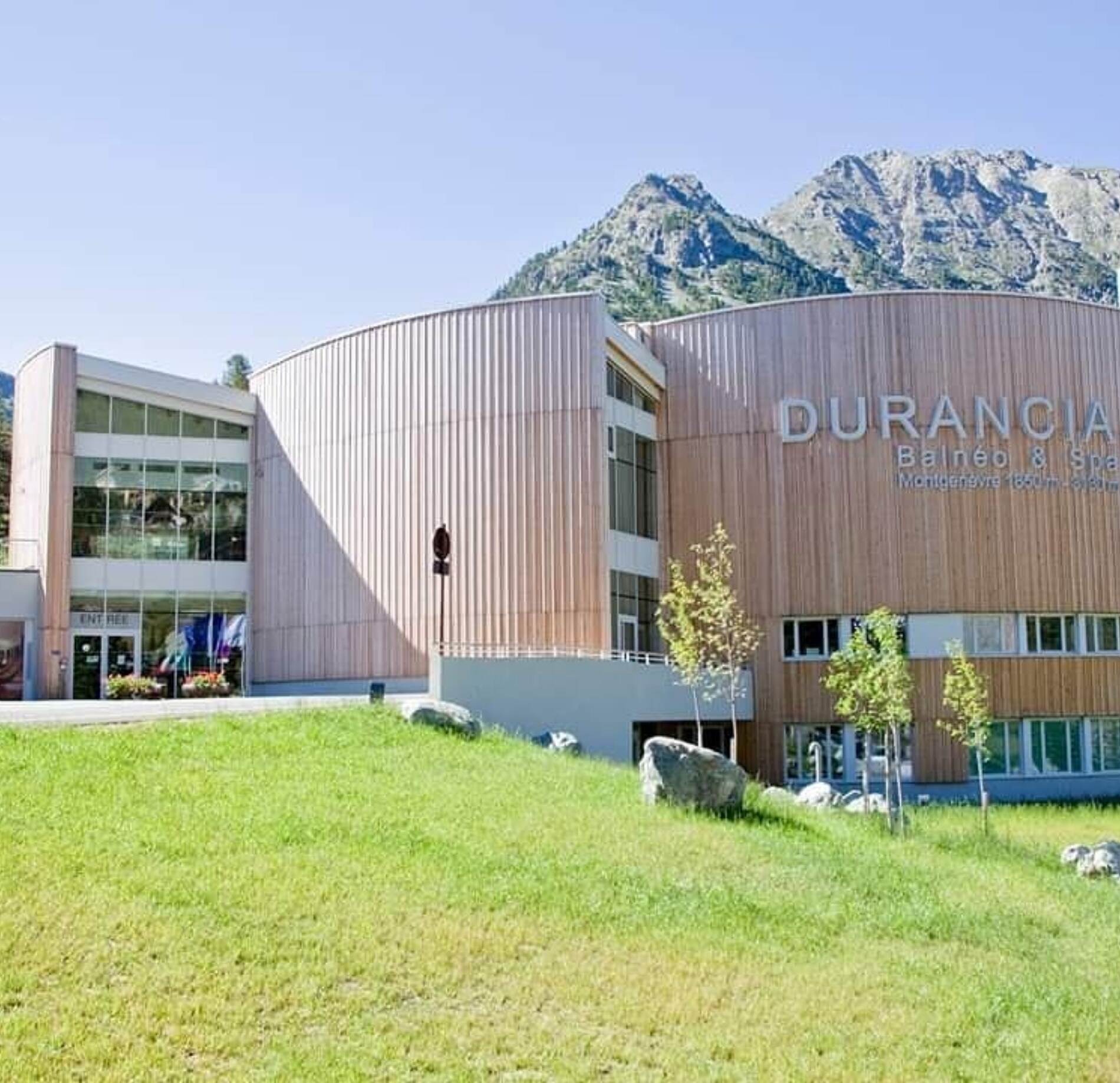 Façade contemporaine du spa Durancia à Montgenèvre, aux lignes arrondies et habillage en bois élégant, située au cœur d’un paysage alpin verdoyant avec montagnes en arrière-plan, sous un ciel clair lumineux, offrant une atmosphère naturelle et apaisante. 