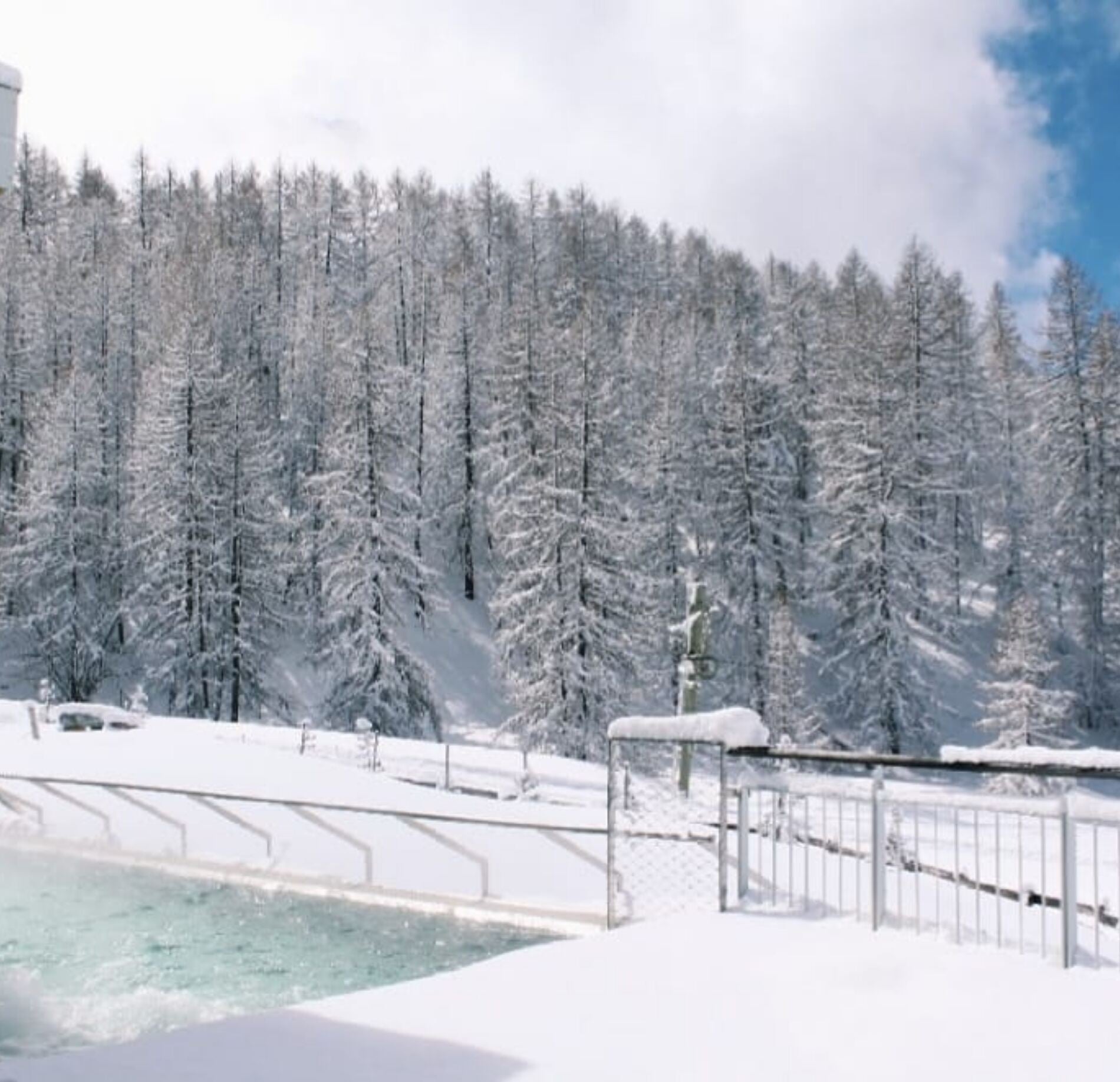 Piscine extérieure élégante entourée d’un manteau de neige immaculé, bordée de sapins enneigés majestueux, avec une eau turquoise contrastant délicatement sous une lumière hivernale douce, créant une atmosphère paisible et ressourçante. 
