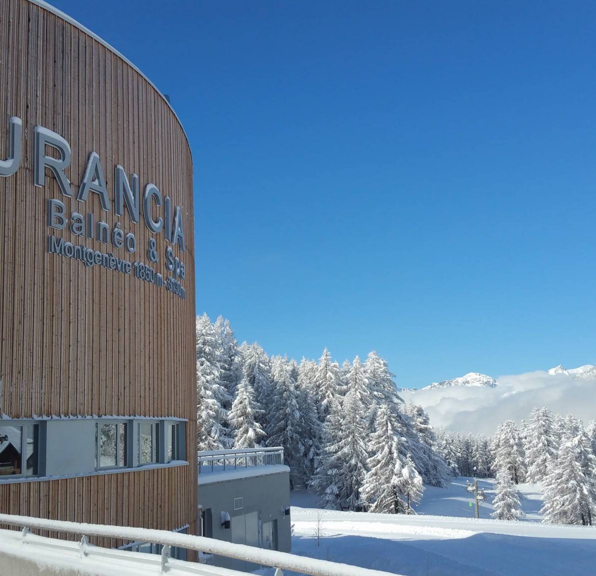 Façade contemporaine du spa Durancia à Montgenèvre, avec bardage en bois élégant, surplombant un paysage montagneux enneigé et des sapins immaculés sous un ciel bleu éclatant, offrant une atmosphère pure et ressourçante