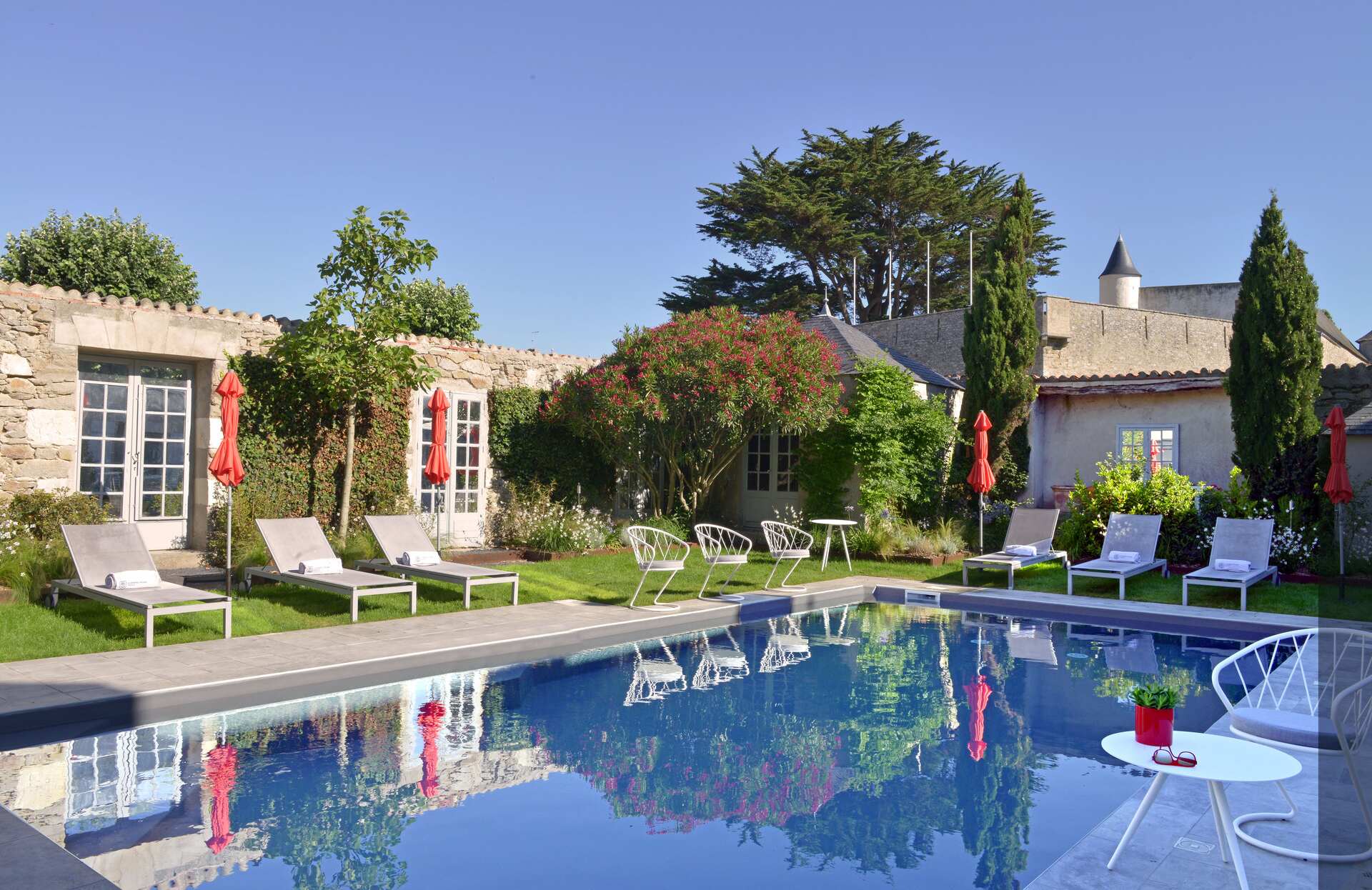 Un charmant espace extérieur au cœur d’un jardin verdoyant, où une piscine aux reflets limpides est entourée de transats élégants et de parasols colorés. Entouré de pierres et de végétation fleurie, ce lieu paisible invite à la détente dans une atmosphère conviviale et authentique.