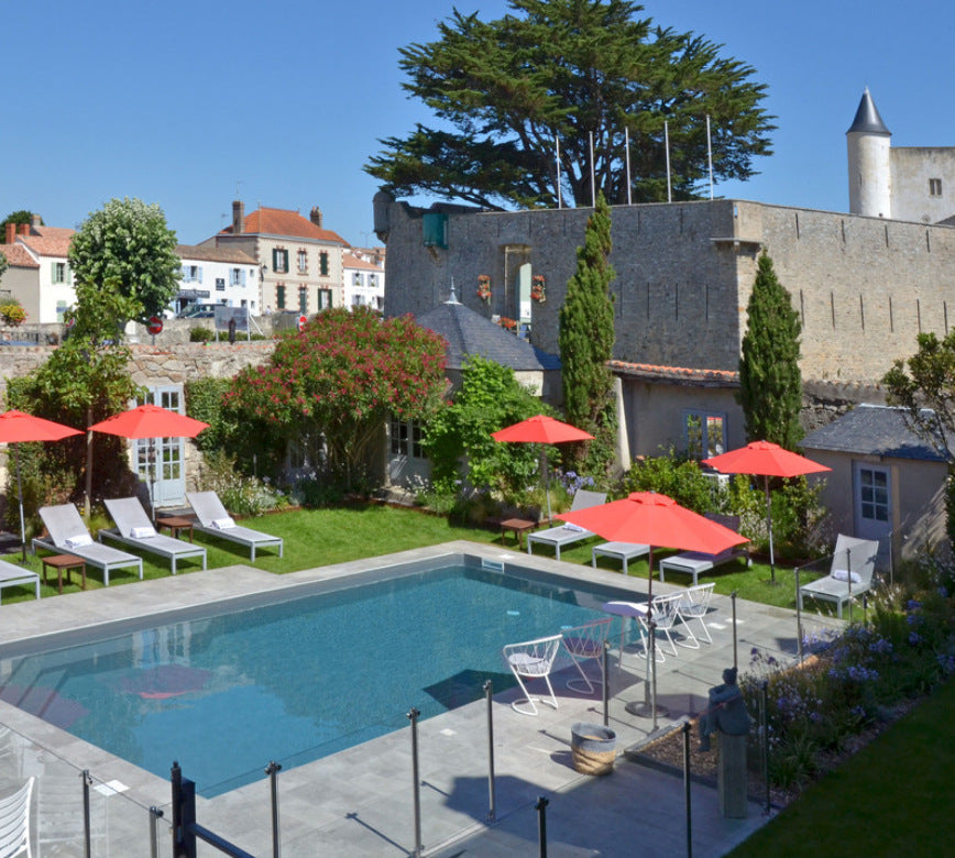 Espace extérieur du spa NUXE à l’Hôtel Elbée, avec bassin piscine aux lignes épurées, terrasse aménagée de transats et parasols colorés, végétation florissante et architecture ancienne en pierre avec remparts, offrant un cadre authentique, paisible et haut de gamme.
