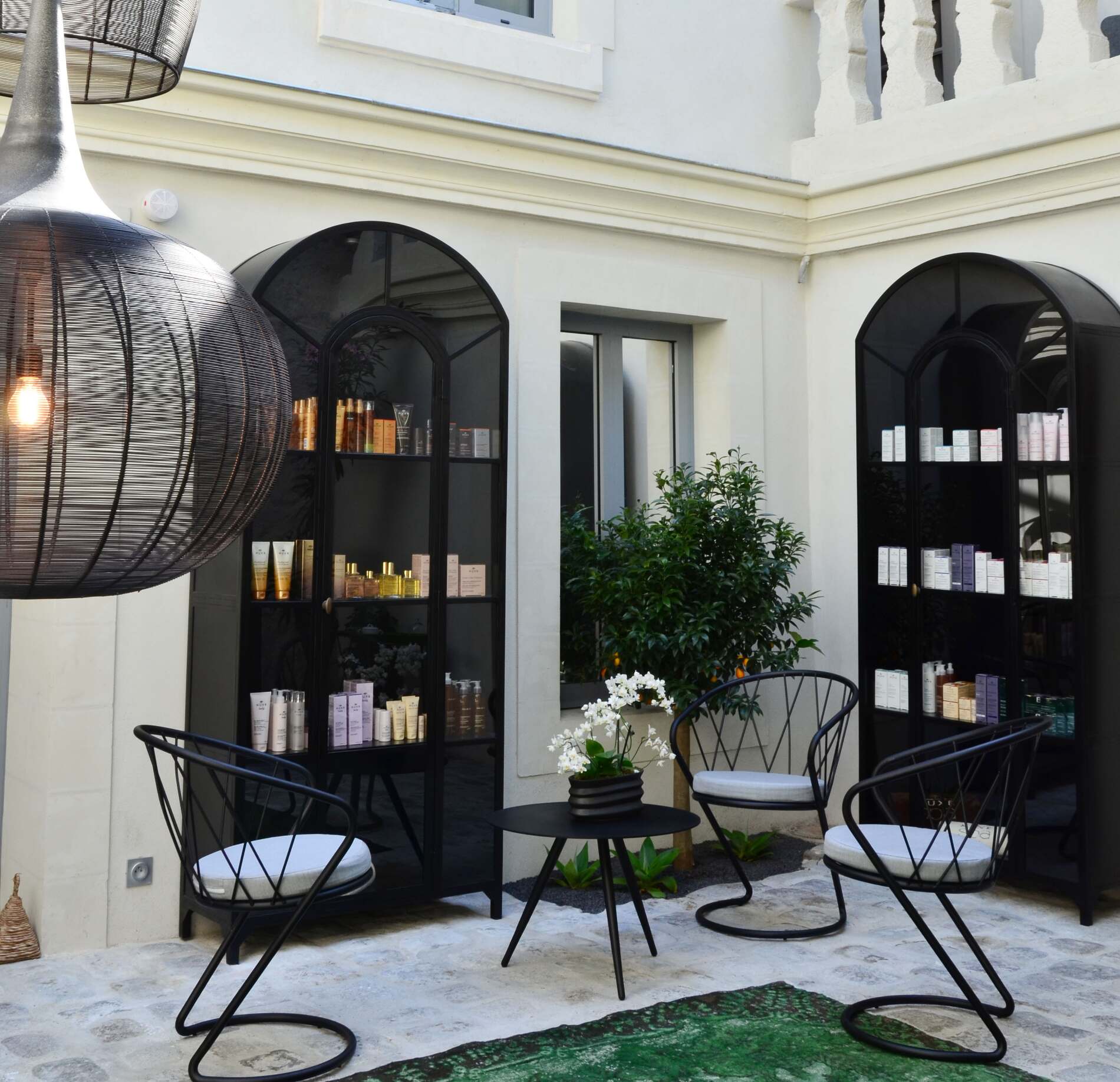 Espace boutique élégant du spa NUXE à l’Hôtel Elbée, aménagé dans une cour intérieure lumineuse avec fauteuils design, table basse raffinée, plantes délicates et étagères présentant des produits de soin, créant une atmosphère chic, apaisante et accueillante. 