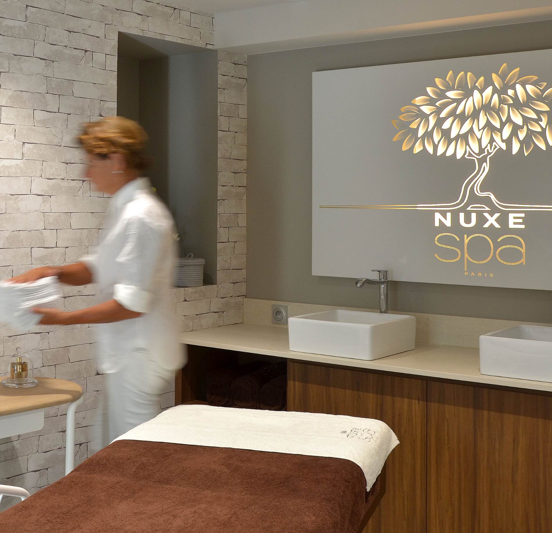 Cabine de soin élégante du spa NUXE à l’Hôtel Elbée, avec table de massage habillée de linge brun, espace lavabo moderne et miroir orné du logo NUXE lumineux, dans une ambiance contemporaine, raffinée et apaisante.