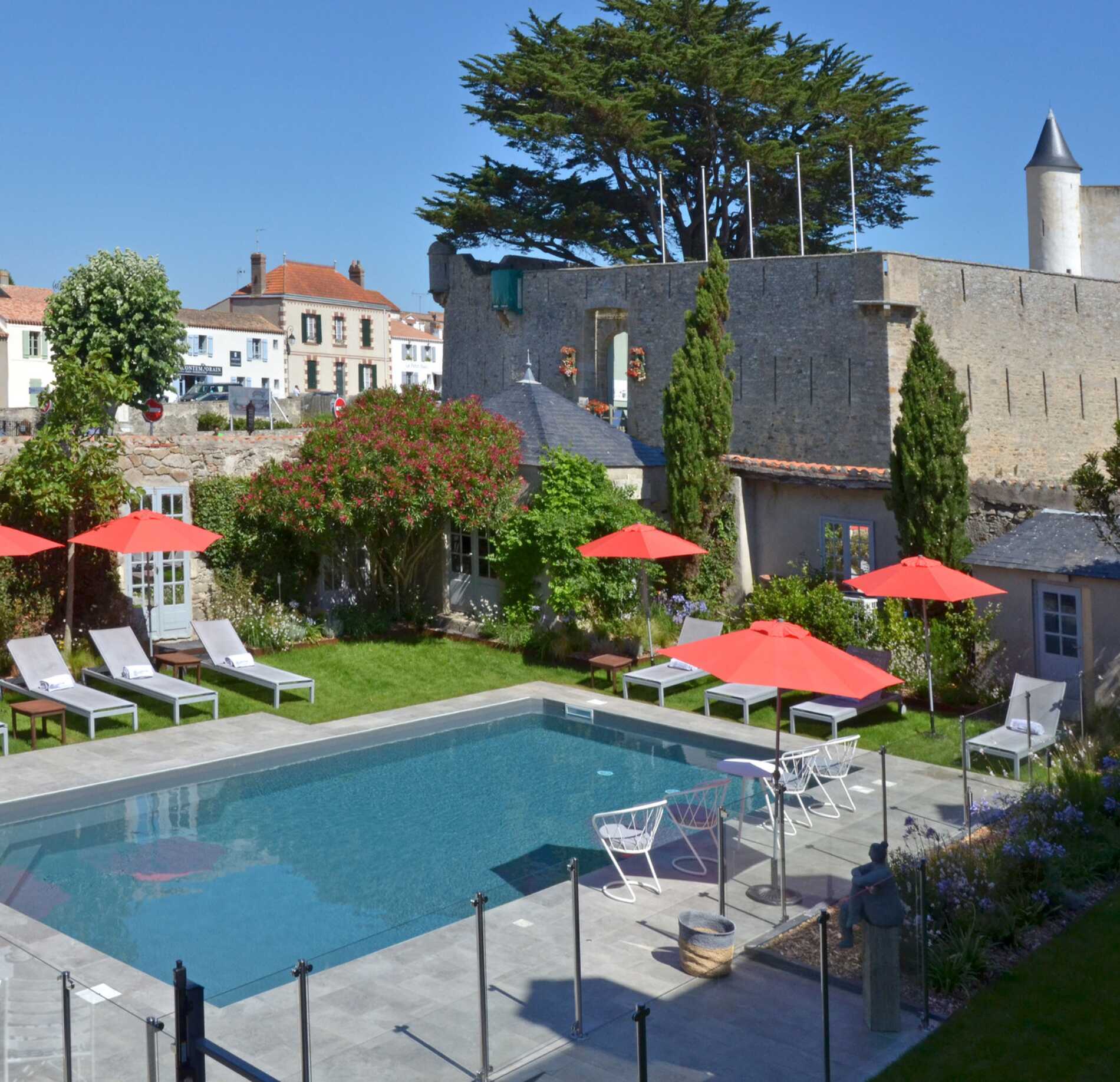 Piscine extérieure élégante du spa NUXE à l’Hôtel Elbée, entourée d’un jardin arboré avec transats et parasols rouges, au pied d’une bâtisse historique en pierre, sous un ciel lumineux créant une atmosphère charmante et raffinée. 