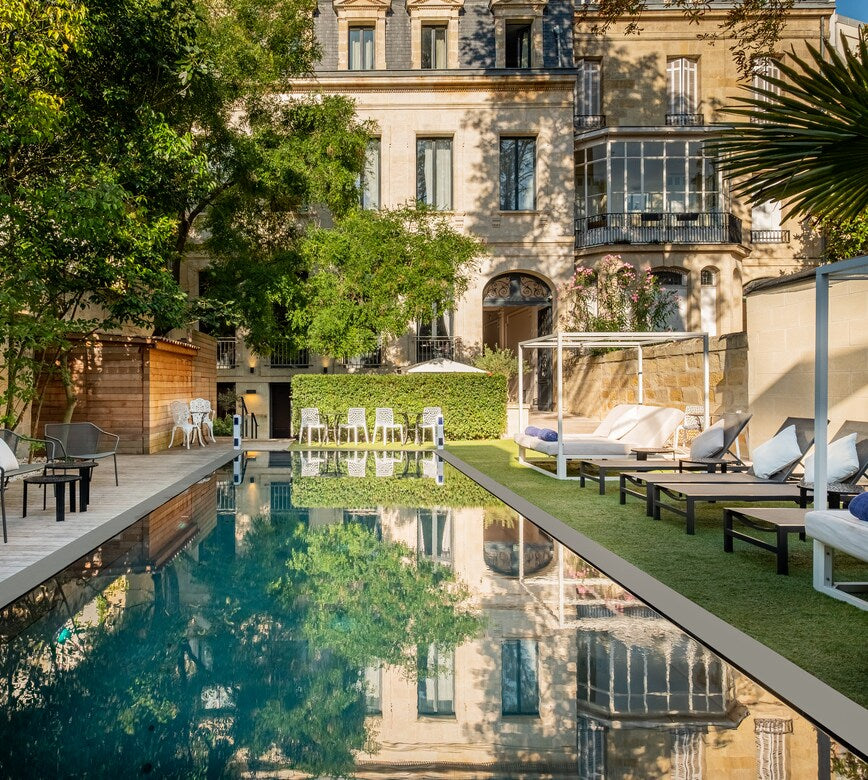 Une piscine extérieure du spa le Palais Gallien s’étend au, entourée de transats et de lits de repos installés sur une terrasse aménagée.
En arrière-plan, un élégant bâtiment ancien avec balcons et grandes fenêtres se dresse au milieu de verdure, se reflétant dans l’eau calme du bassin.