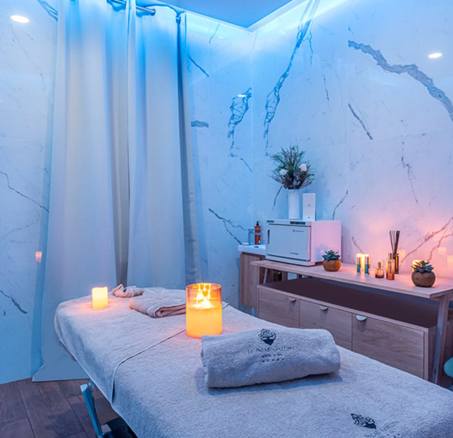 Une cabine de soins du spa le Palais Gallien présente une table de massage recouverte de linges clairs, avec une serviette roulée et des bougies allumées posées dessus.
Autour, des murs effet marbre, un meuble en bois avec produits de soin et une lumière douce bleutée créent une ambiance apaisante et intime.