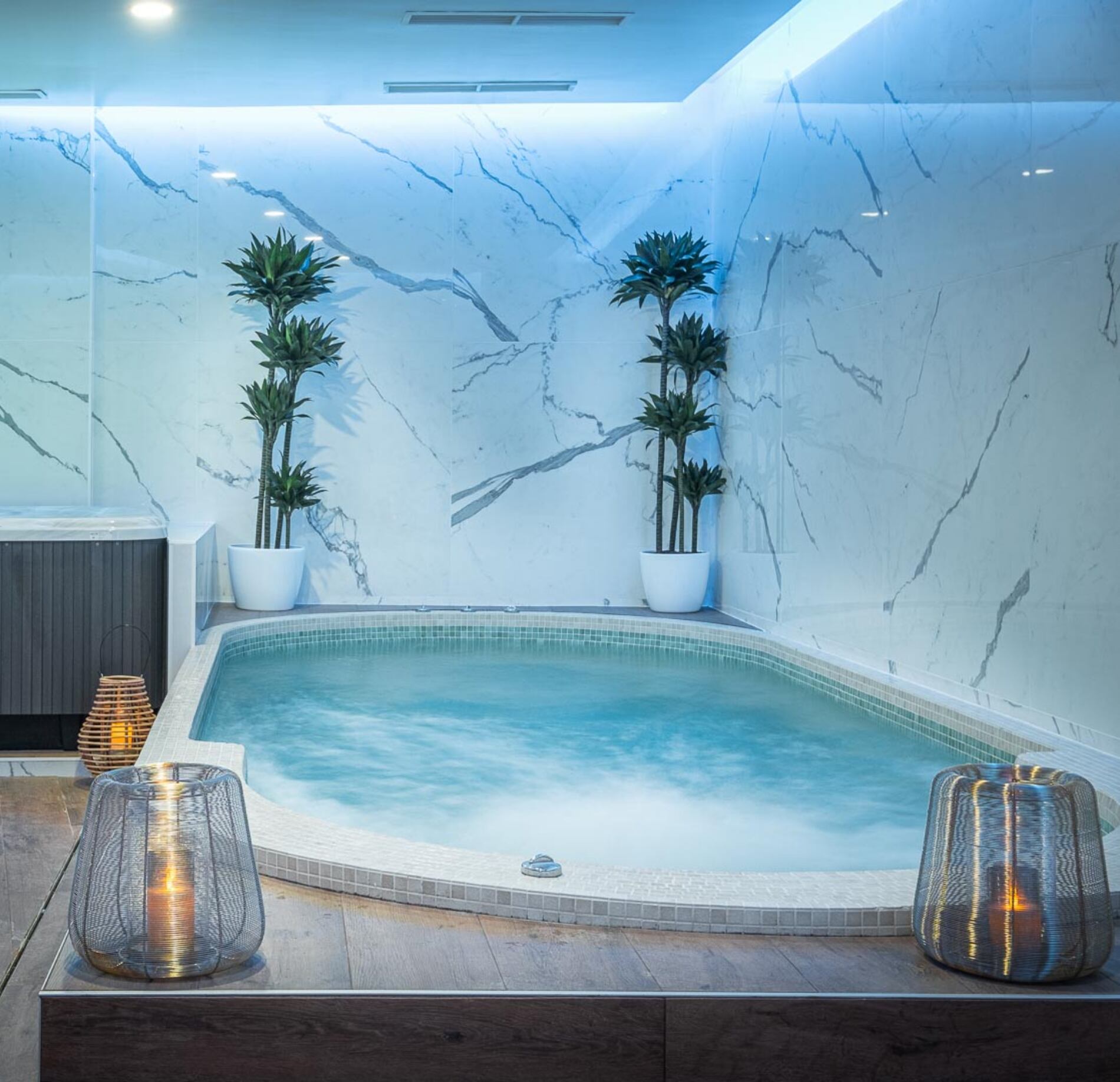 Un jacuzzi du spa le Palais Gallien est encastré dans le sol, avec une eau claire en mouvement entourée de carrelage clair et d’un rebord arrondi.
Autour, des murs effet marbre, des plantes en pots et des bougies dans des lanternes créent une ambiance douce, moderne et relaxante.