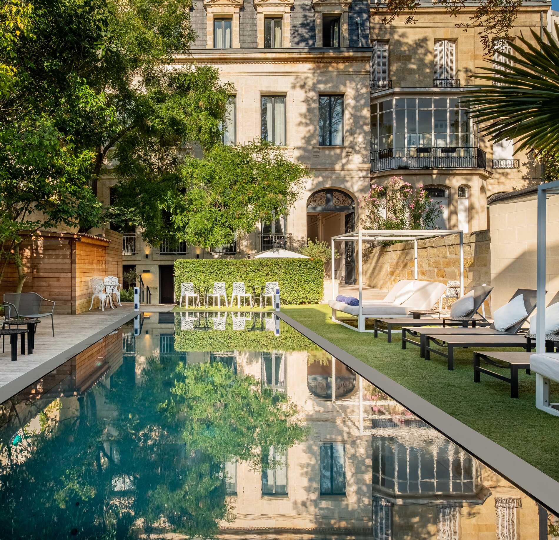 Une piscine extérieure du spa le Palais Gallien s’étend au premier plan, entourée de transats et de lits de repos installés sur une terrasse aménagée.
En arrière-plan, un élégant bâtiment ancien avec balcons et grandes fenêtres se dresse au milieu de verdure, se reflétant dans l’eau calme du bassin.