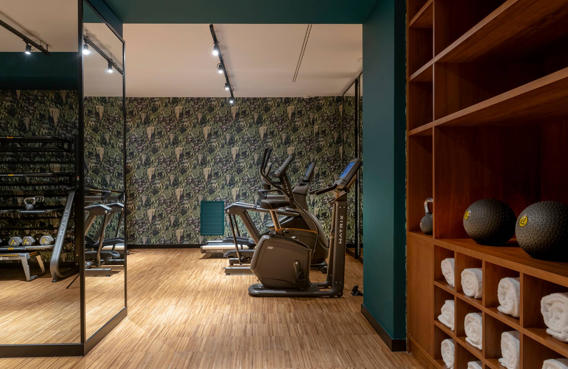 Une salle de sport moderne et parfaitement équipée, avec des machines de cardio alignées sur un parquet chaleureux et un mur végétal décoratif apportant une touche naturelle.
Les rangements en bois, les serviettes soigneusement disposées et l’éclairage ciblé créent une atmosphère haut de gamme, propice à une expérience bien‑être complète et dynamique.
