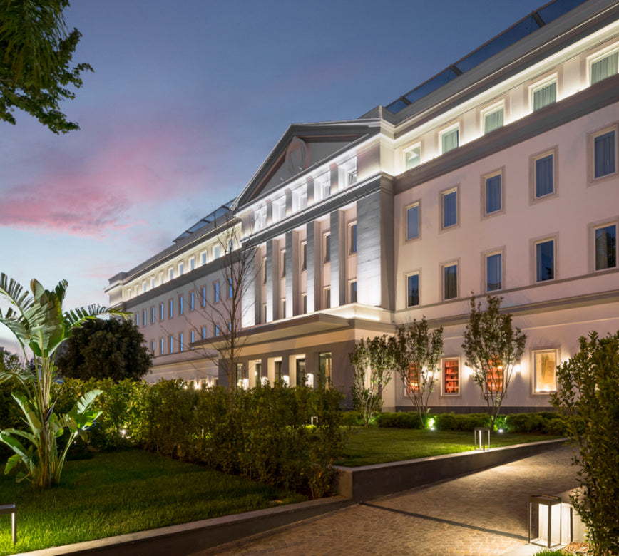 Façade majestueuse d’un établissement de standing, illuminée par un éclairage architectural chaleureux, avec grandes fenêtres régulières, jardin paysager et allées discrètes, dans une ambiance paisible et haut de gamme au coucher du jour. 