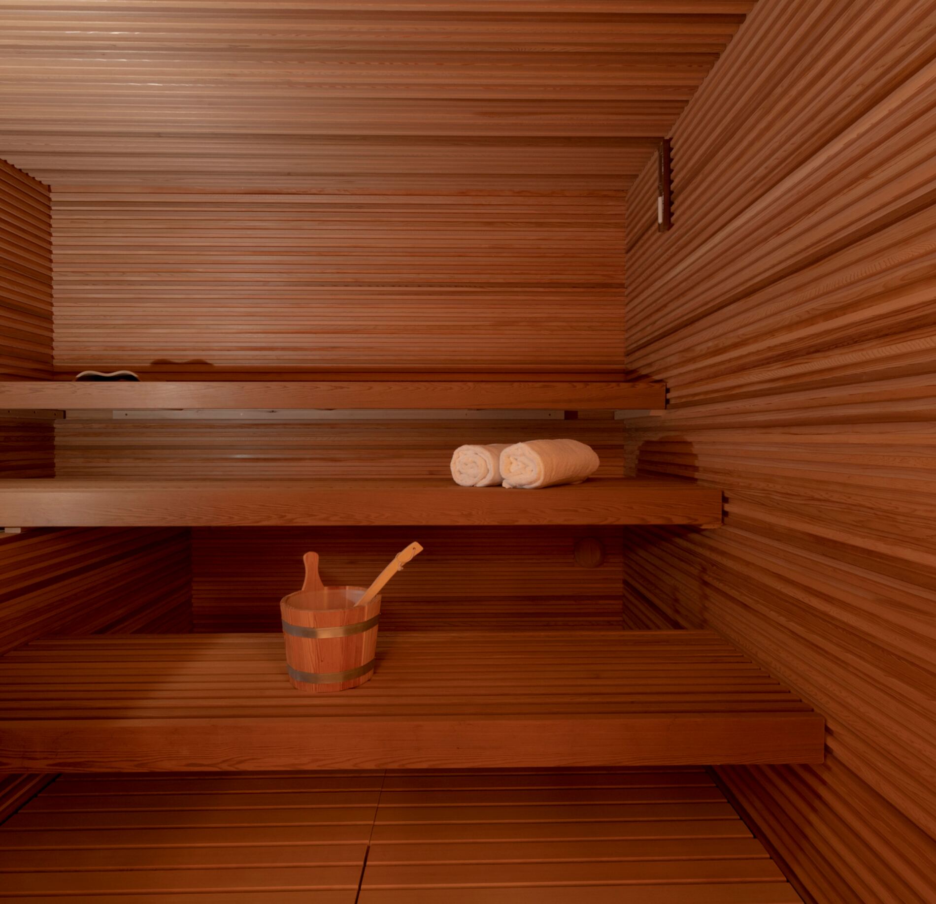 Sauna élégant aux parois en bois chaleureux, équipé de banquettes spacieuses et d’accessoires traditionnels, baigné d’une lumière douce créant une atmosphère intime et profondément relaxante. 