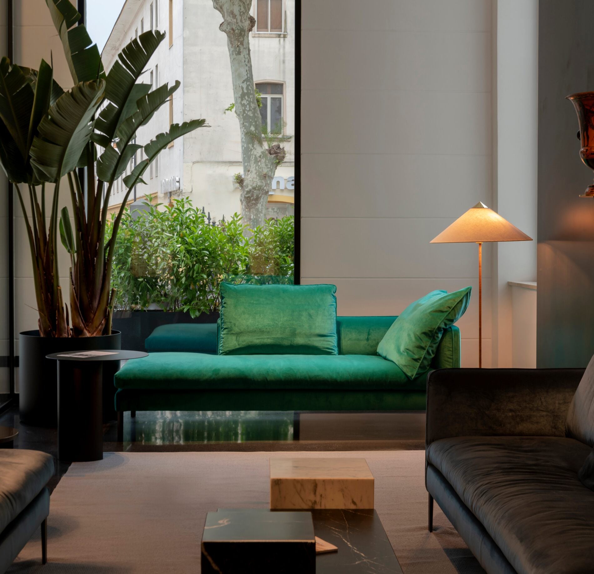 Espace lounge élégant et contemporain, avec canapé vert profond aux coussins moelleux, éclairé par une lampe design chaleureuse, entouré de plantes verdoyantes et baigné de lumière naturelle pour une atmosphère raffinée et apaisante.