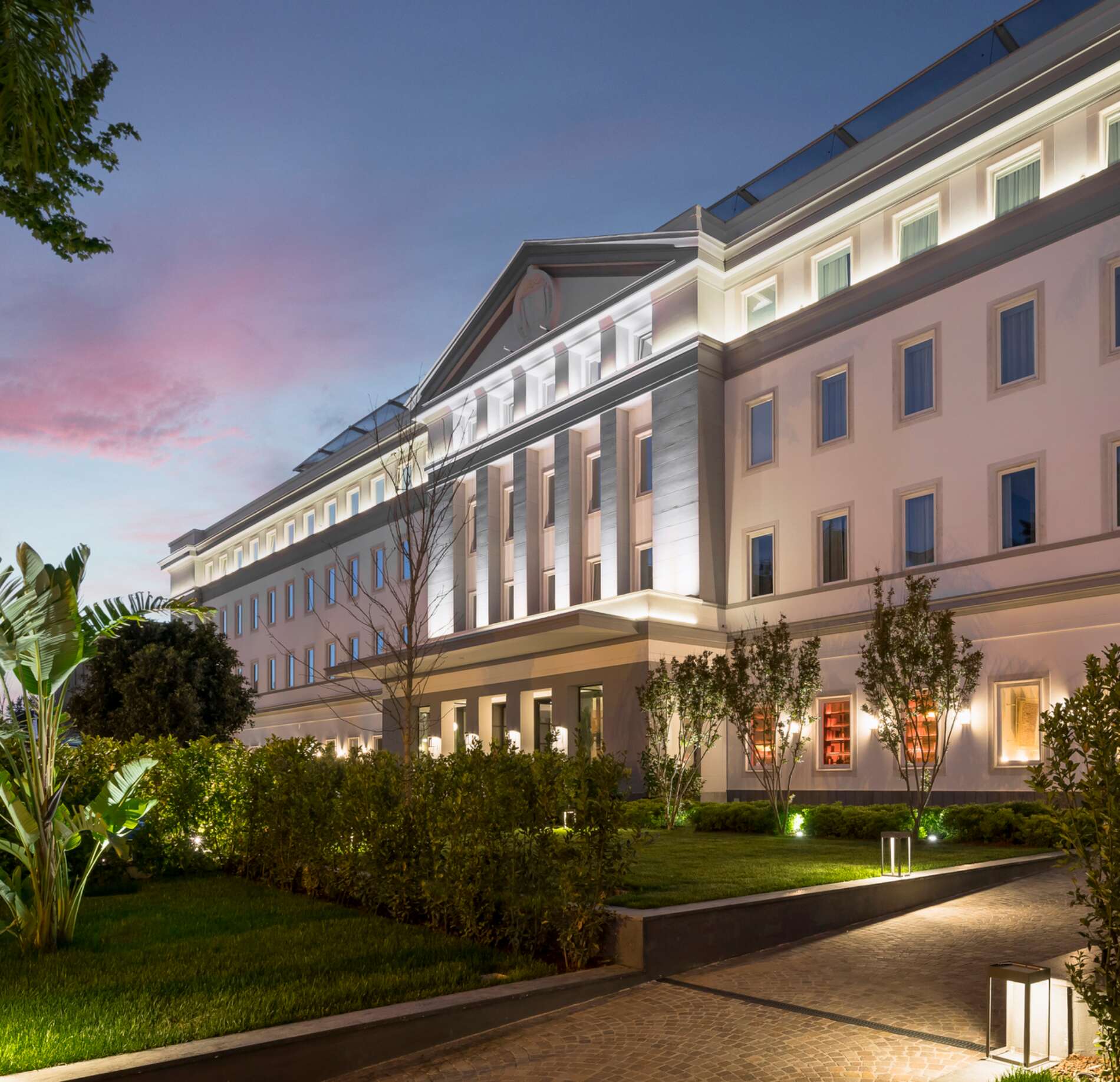 Élégant bâtiment hôtelier aux lignes classiques, subtilement éclairé en soirée, entouré d’un jardin soigné et de verdure, sous un ciel aux teintes douces du crépuscule, créant une atmosphère raffinée et accueillante.