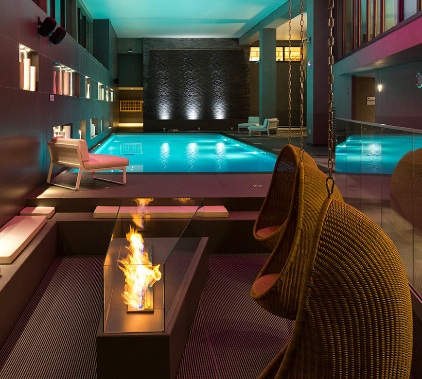 Espace aquatique contemporain du spa NUXE à l’Hôtel Héliopic, avec grand bassin intérieur aux reflets lumineux, éclairages d’ambiance colorés, fauteuils confortables, balancelles suspendues en fibres naturelles et cheminée décorative, offrant un cadre immersif, convivial et haut de gamme propice à la détente.