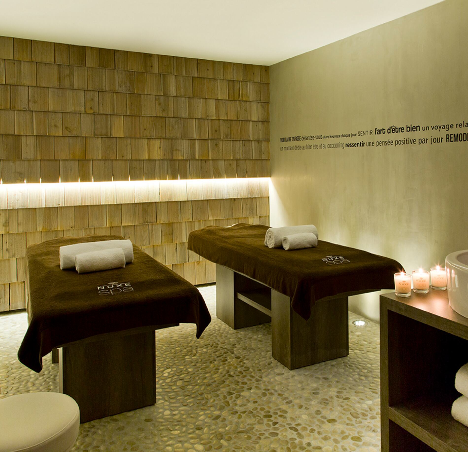 Cabine de soin chaleureuse du spa NUXE à l’Hôtel Héliopic, avec deux tables de massage habillées de linge brun, éclairage doux, mur en bois texturé et bougies disposées avec soin, créant une atmosphère apaisante, élégante et haut de gamme.
