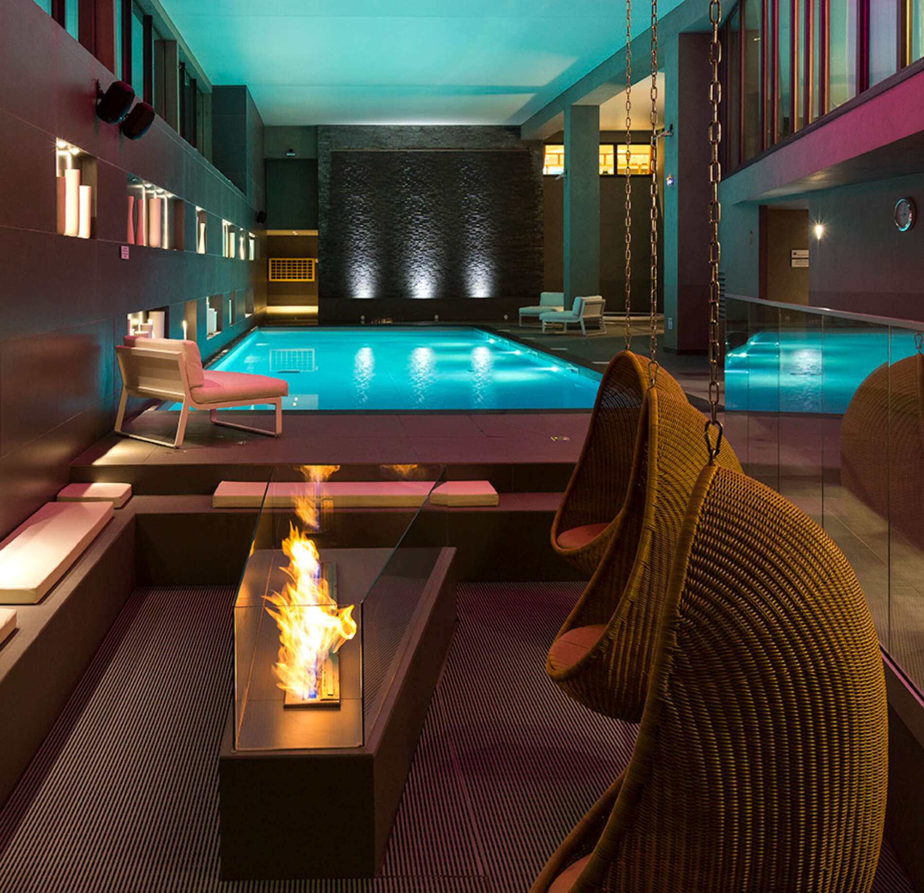 Piscine intérieure élégante du spa NUXE à l’Hôtel Héliopic, baignée de lumières colorées turquoise et ambre, avec bassin apaisant, espace détente raffiné, fauteuils design et coin cheminée moderne créant une atmosphère chaleureuse et contemporaine. 