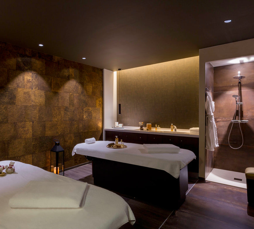 Salle de massage contemporaine du spa NUXE à l’Hôtel de la Poste Troyes, composée de tables de soin soigneusement préparées, d’un mur à l’aspect minéral chaleureux, d’éclairages doux et d’une cabine de douche ouverte, offrant un cadre serein, haut de gamme et propice à la détente.
