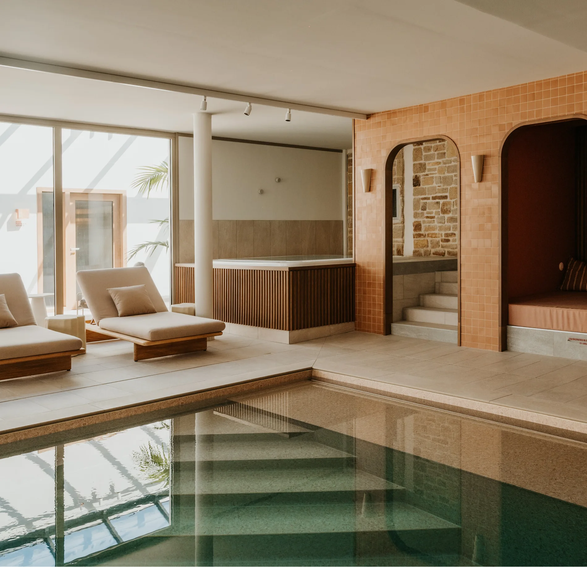 Espace spa intérieur avec bassin, chaises longues et bain à remous, dans un environnement lumineux aux murs en pierre et en carrelage.