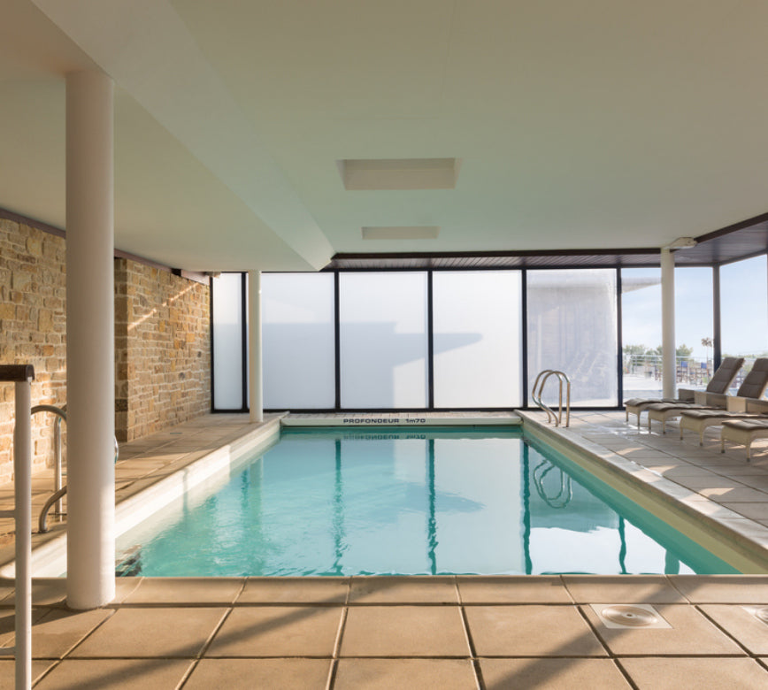 Une piscine intérieure lumineuse du Spa Hostellerie de la Pointe Saint Mathieu, avec une eau turquoise bordée de larges baies vitrées ouvrant sur l’extérieur.
Les murs mêlant pierre et surfaces claires, ainsi que des transats alignés au fond, créent une atmosphère apaisante et élégante propice à la détente.