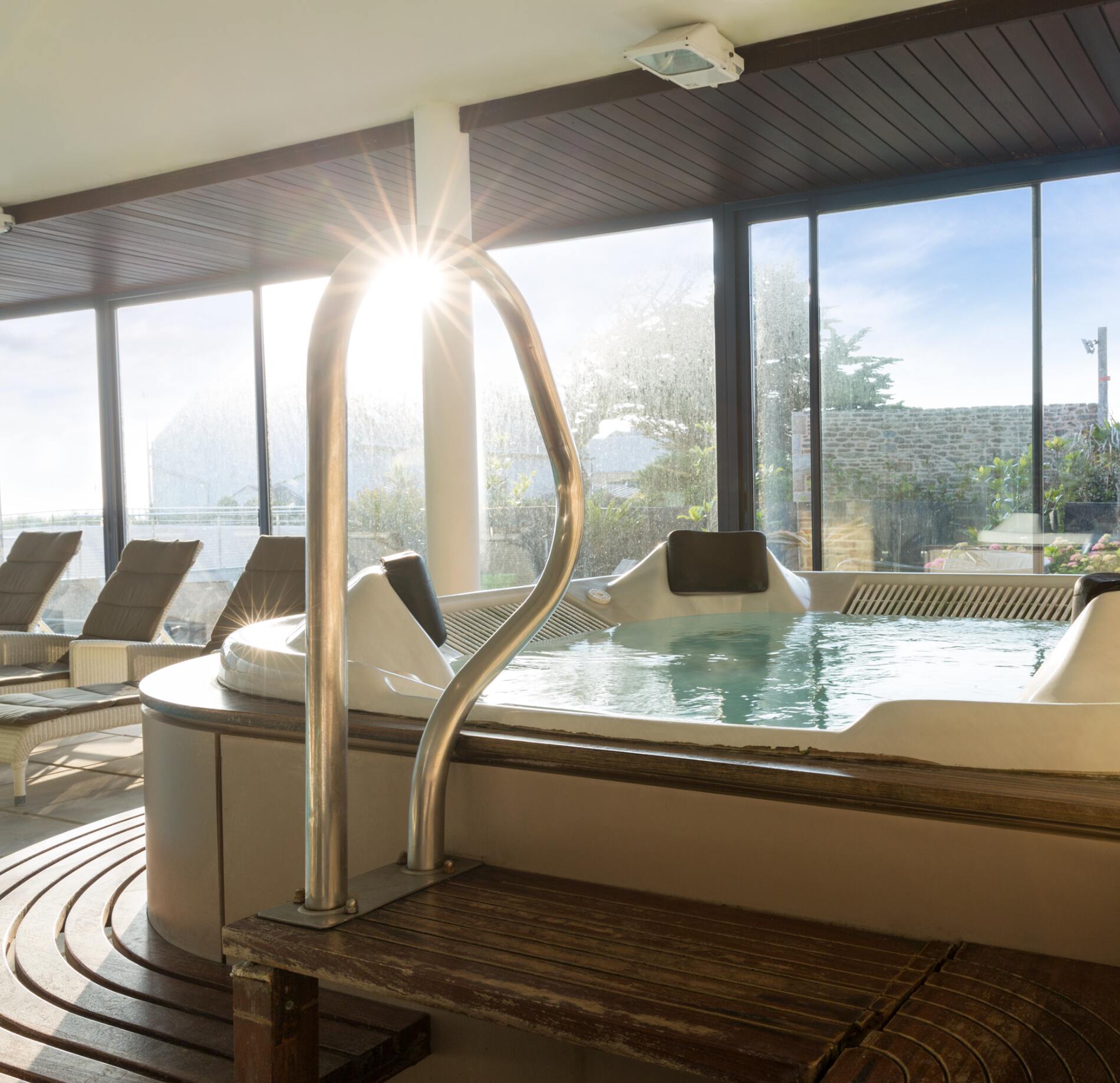 Un jacuzzi lumineux du Spa Hostellerie de la Pointe Saint Mathieu, avec une eau légèrement agitée entourée de grandes baies vitrées laissant entrer la lumière du soleil.
Au premier plan, une rampe métallique et un plancher en bois humide reflètent la lumière, tandis que des transats alignés complètent cette atmosphère apaisante et élégante.