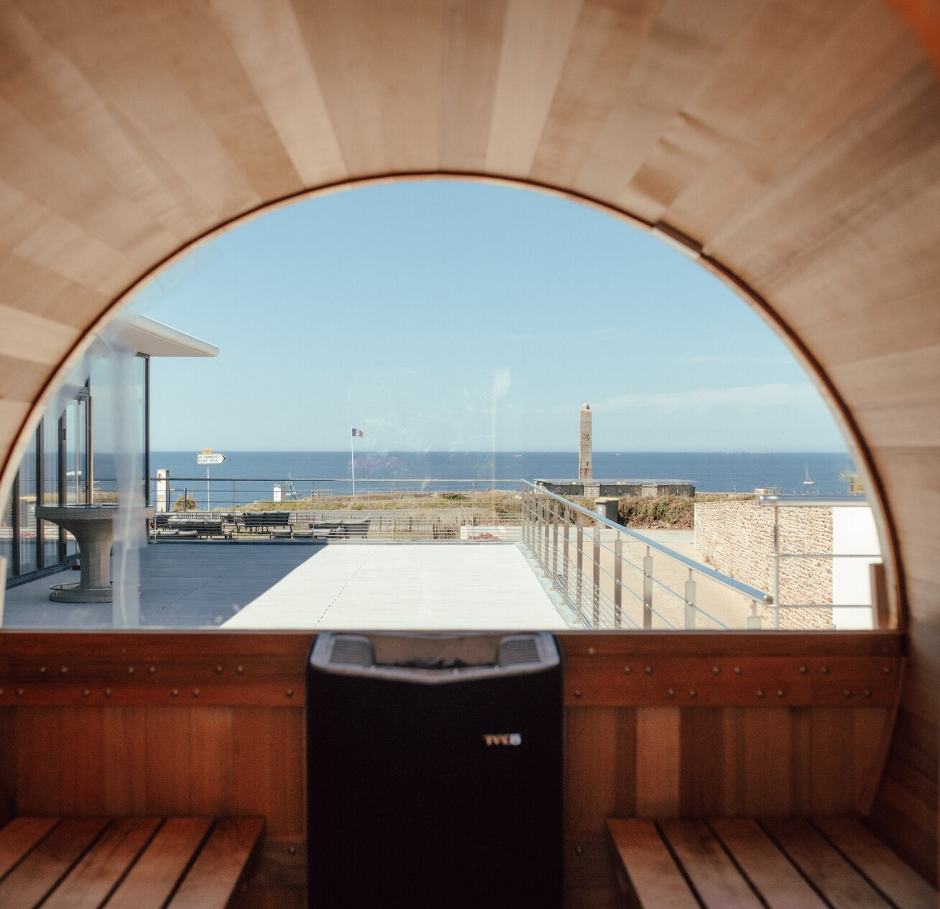 Un sauna avec vue ouverte du Spa Hostellerie de la Pointe Saint Mathieu, encadré par une large ouverture arrondie donnant sur une terrasse et l’océan à l’horizon.
Les bancs en bois clair et les parois chaleureuses contrastent avec le bleu du ciel et de la mer, offrant une atmosphère apaisante entre intérieur et extérieur.