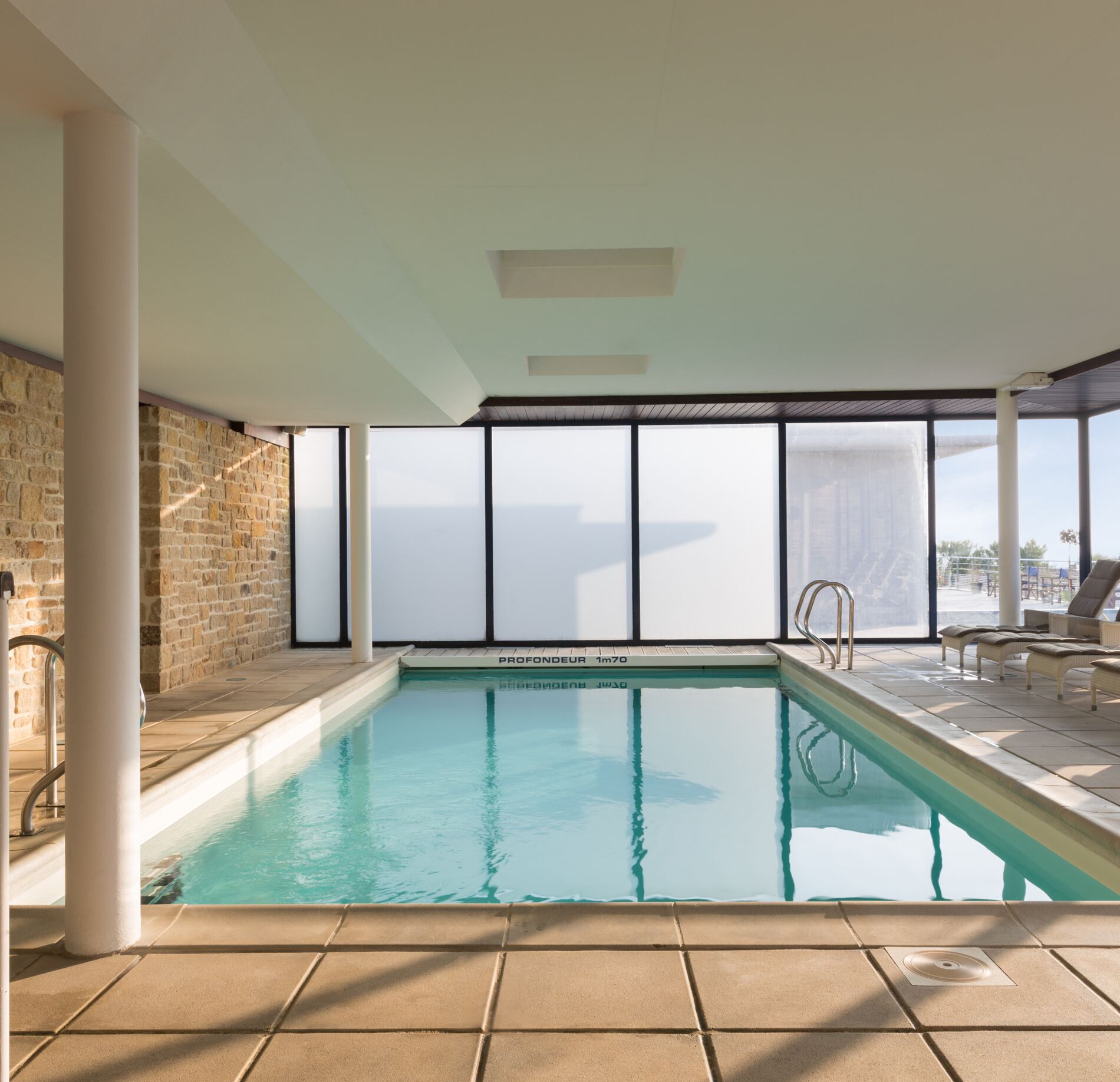 Une piscine intérieure lumineuse du Spa Hostellerie de la Pointe Saint Mathieu, avec une eau turquoise entourée de grandes baies vitrées ouvertes sur l’extérieur.
Les murs associant pierre et surfaces claires, ainsi que des transats disposés au fond, créent une ambiance sereine et raffinée idéale pour la détente.
