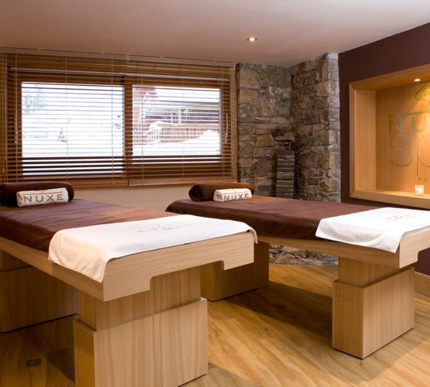 Une salle de soins du spa Nuxe de l'hotel le Kaïla présente deux tables de massage en bois clair, recouvertes de linges blancs et de serviettes roulées brodées NUXE.
En arrière-plan, un mur en pierre et une fenêtre avec des stores laissent entrer la lumière, créant une atmosphère calme et chaleureuse propice à la détente.