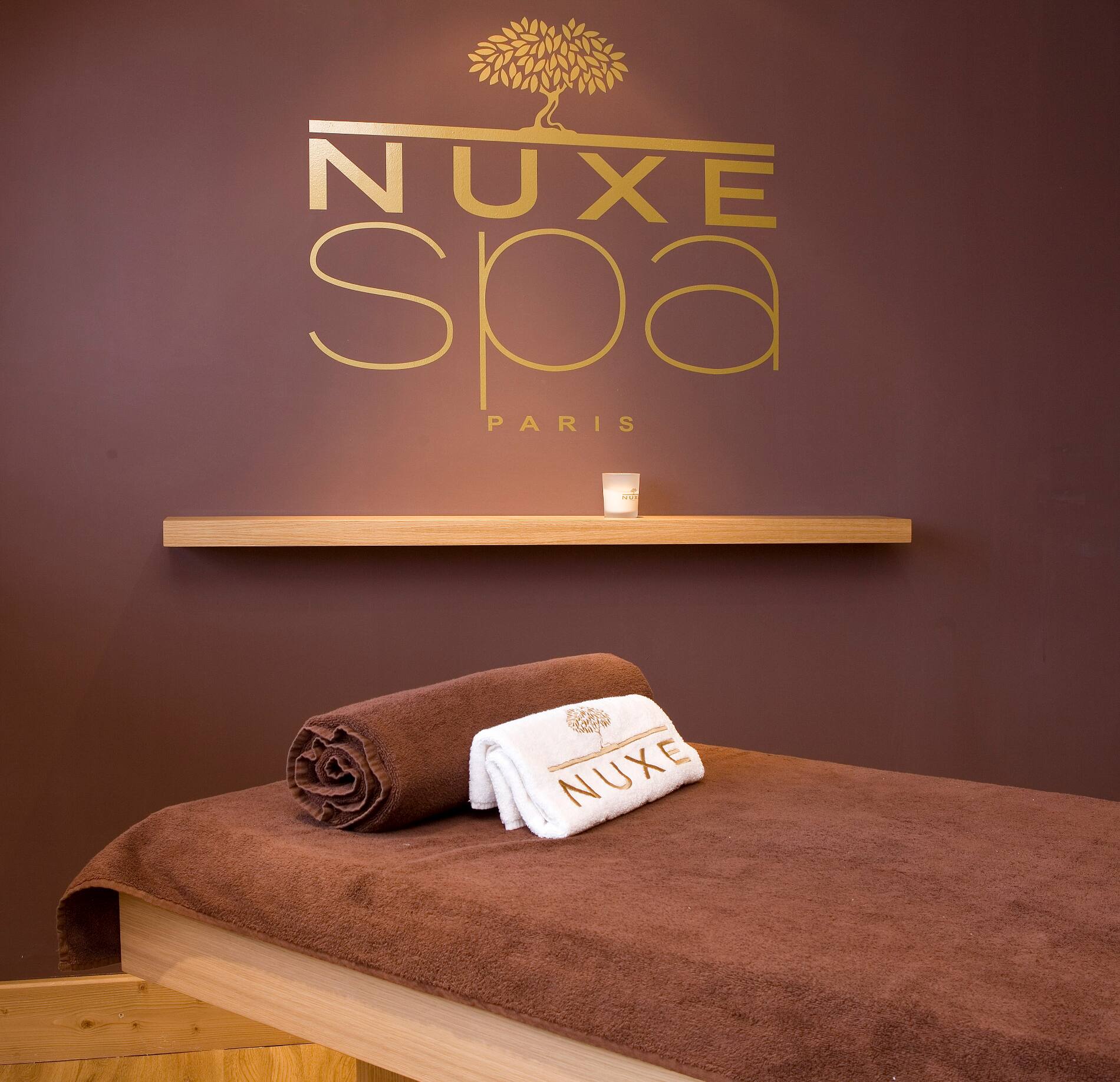 Une table de soin du spa Nuxe de l'hotel le Kaïla est recouverte d’une serviette marron avec deux serviettes roulées, dont une brodée “NUXE”, posées au centre.
Au mur, le logo doré “NUXE Spa Paris” se détache sur un fond brun, avec une petite étagère en bois et une bougie créant une ambiance chaleureuse et apaisante.