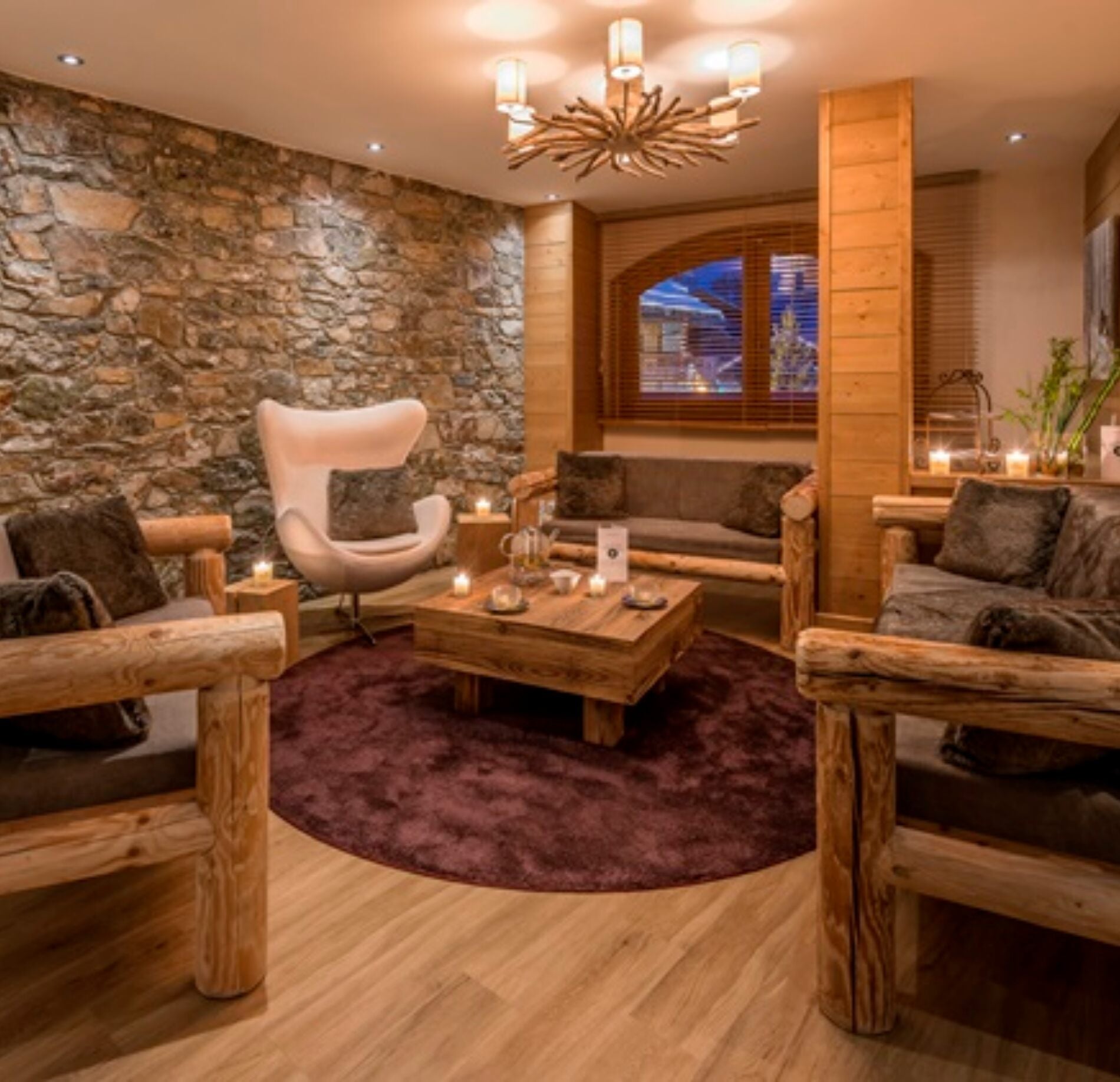 Un salon de détente du spa Nuxe de l'hotel le Kaïla est aménagé avec des fauteuils et canapés en bois massif disposés autour d’une table basse, sur un tapis moelleux.
Un mur en pierre, des bougies et un éclairage chaleureux créent une atmosphère cosy, tandis qu’une grande fenêtre laisse entrevoir l’extérieur.