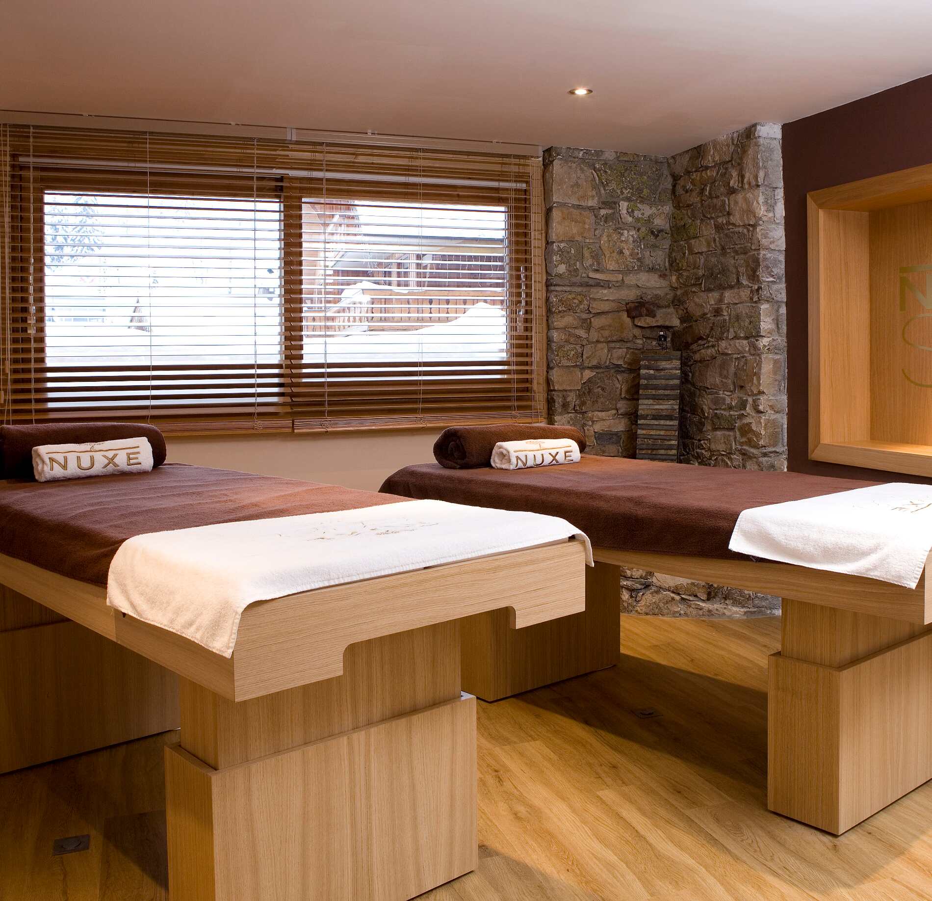 Une salle de soins du spa Nuxe de l'hotel le Kaïla présente deux tables de massage en bois clair, recouvertes de linges blancs et de serviettes roulées.
En arrière-plan, un mur en pierre et une fenêtre avec des stores laissent entrer la lumière, créant une atmosphère calme et chaleureuse propice à la détente.