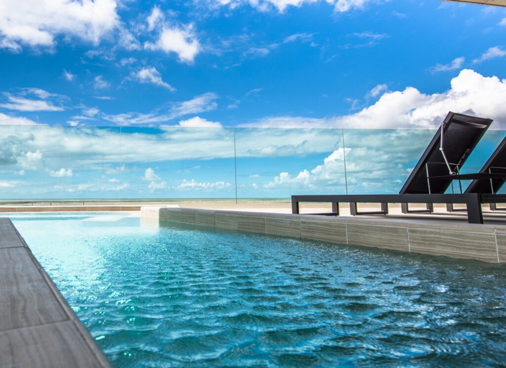 Piscine moderne avec vue sur la mer, terrasse équipée de transats et garde-corps en verre sous un ciel bleu ensoleillé