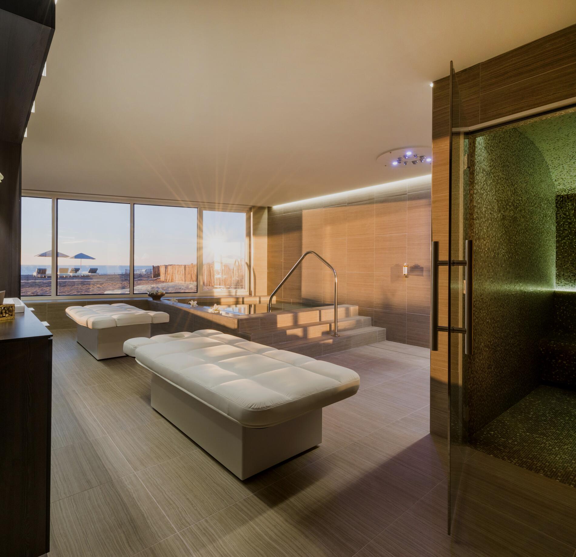 Espace bien-être lumineux du spa NUXE à La Grande Terrasse, avec bancs de repos élégants, bassin chauffé accessible par marches et large baie vitrée offrant une vue apaisante sur l’océan baigné de lumière naturelle.