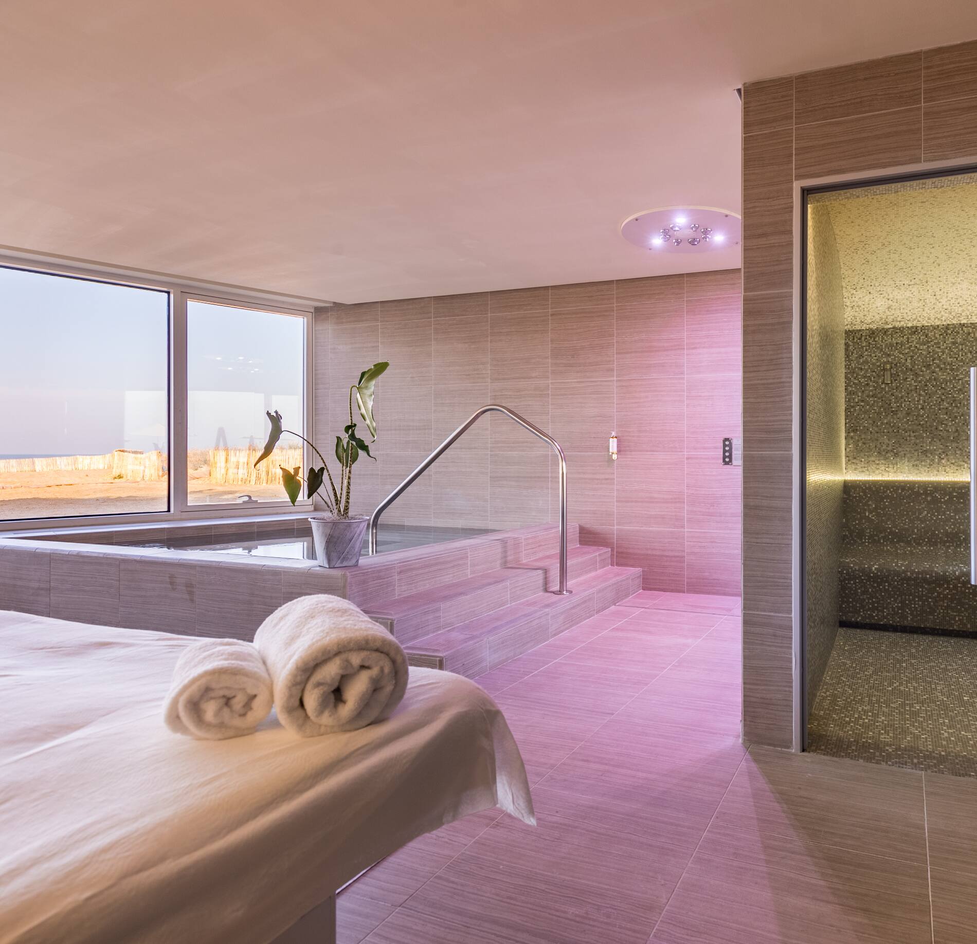 Espace bien-être élégant du spa NUXE à La Grande Terrasse, avec bassin intérieur accessible par marches, banquettes de repos confortables et hammam contemporain, baigné d’une lumière douce rosée et ouvert sur l’extérieur grâce à de larges baies vitrées offrant une vue apaisante sur l’océan. 