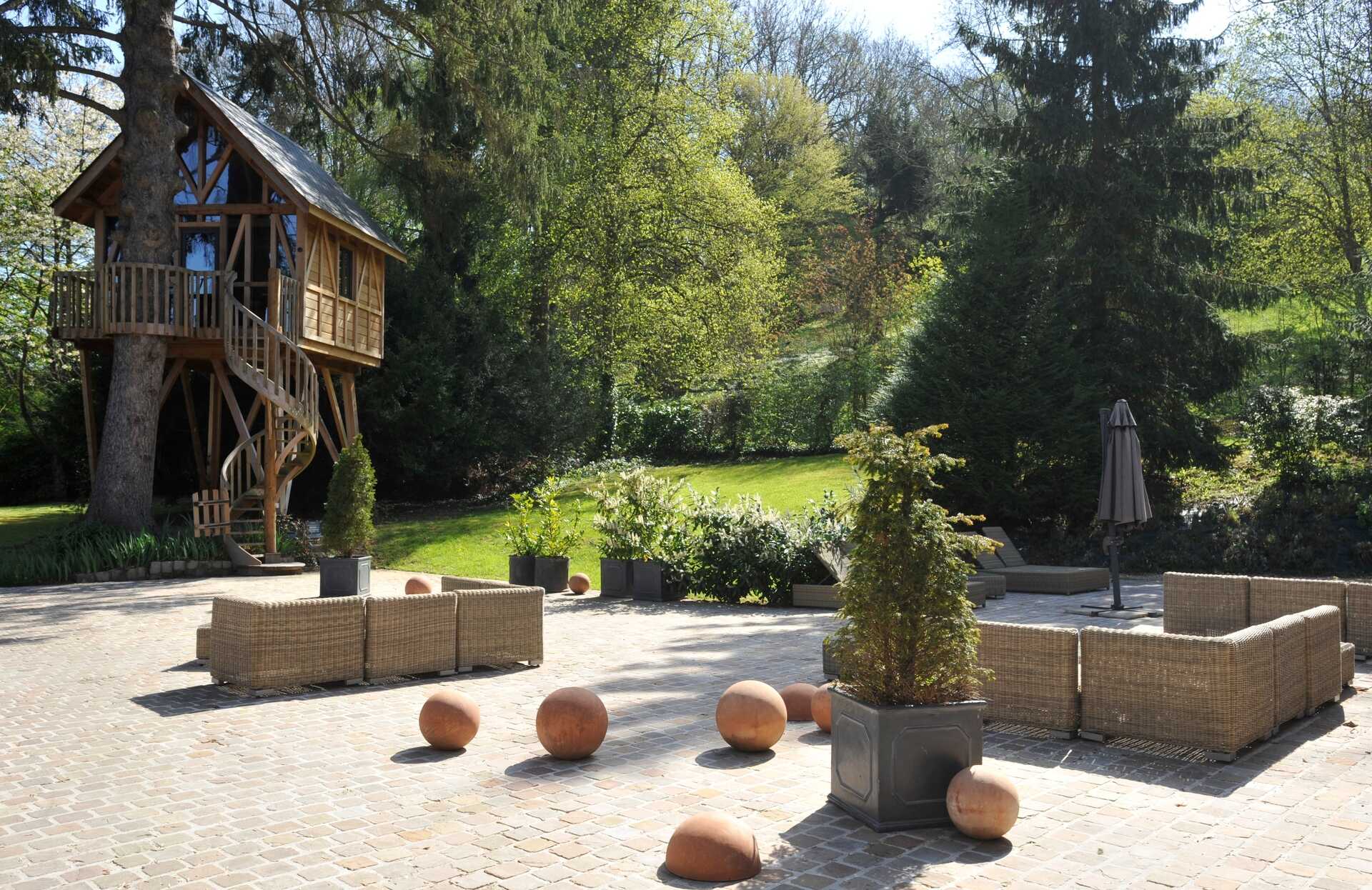 Un espace extérieur paisible s’ouvre sur une terrasse pavée aménagée de salons en rotin, entourée de végétation dense et baignée de lumière naturelle.
Sur la gauche, une cabane perchée en bois accessible par un escalier ajoute une touche unique et immersive, offrant une expérience exceptionnelle au cœur de la nature.