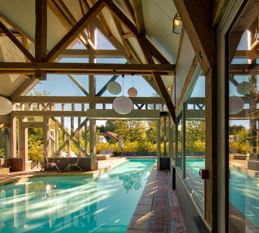 Bassin intérieur du spa NUXE à l’Hôtel La Licorne, niché dans un espace baigné de lumière, avec charpente en bois majestueuse, suspensions contemporaines, larges ouvertures sur l’extérieur et transats disposés en bord de piscine, offrant un cadre chaleureux.