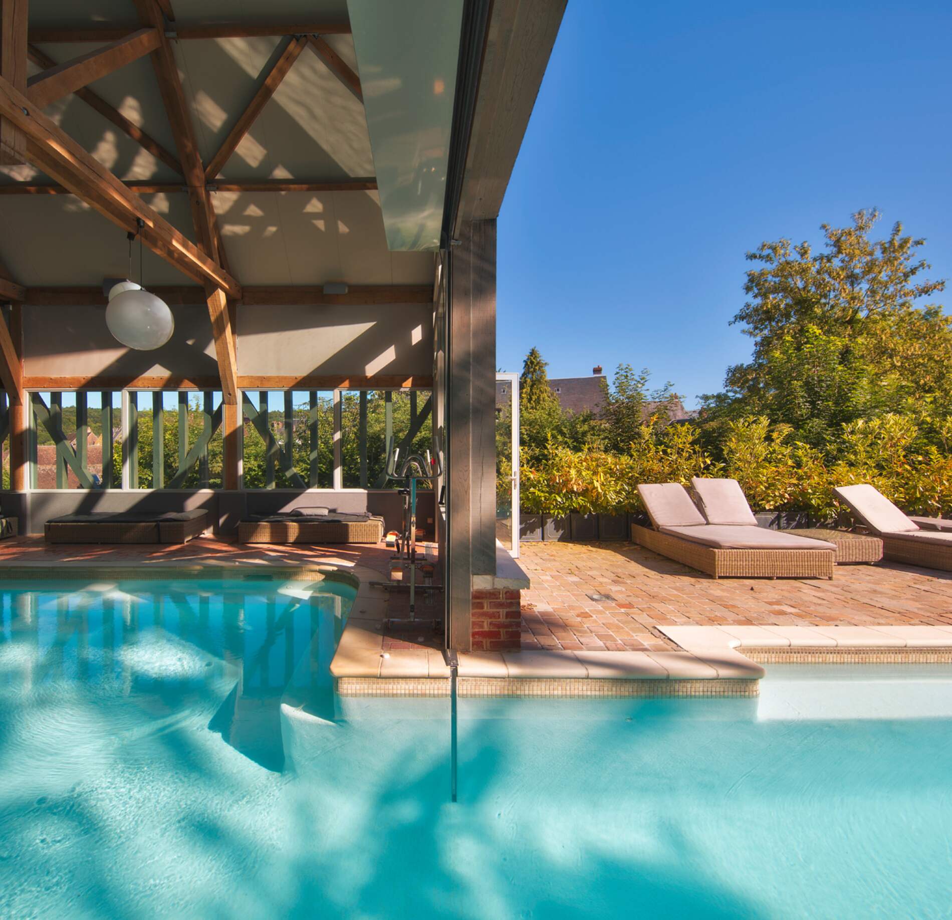 Piscine intérieure-extérieure du spa NUXE à l’Hôtel La Licorne, avec eau turquoise lumineuse, structure en bois apparente et ouverture sur une terrasse ensoleillée équipée de transats, entourée de végétation verdoyante sous un ciel bleu éclatant.