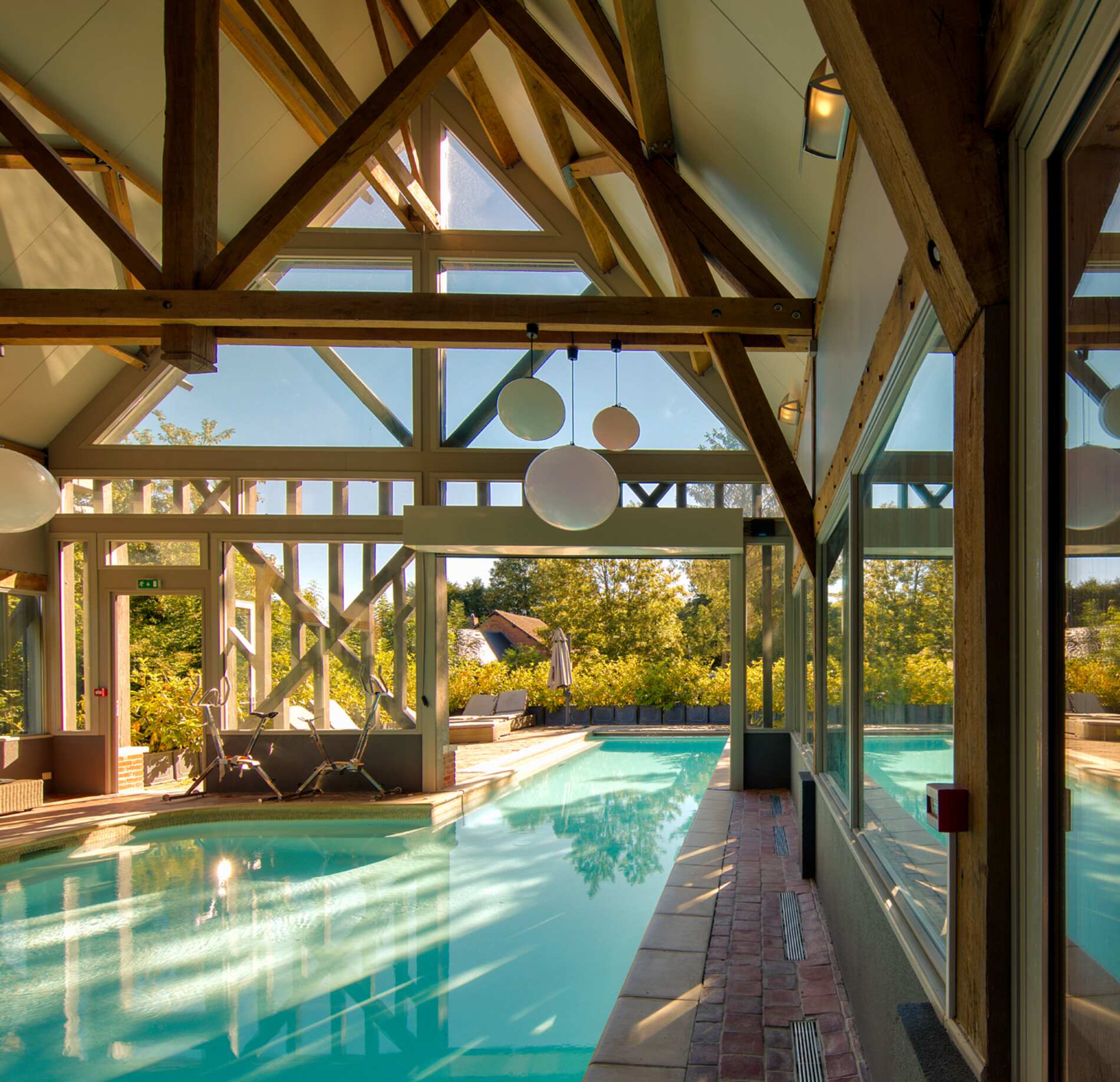 Piscine intérieure lumineuse du spa NUXE à l’Hôtel La Licorne, installée sous une charpente en bois apparente avec grandes baies vitrées, eau turquoise scintillante et vue sur un jardin verdoyant, créant une atmosphère naturelle, élégante et apaisante.
