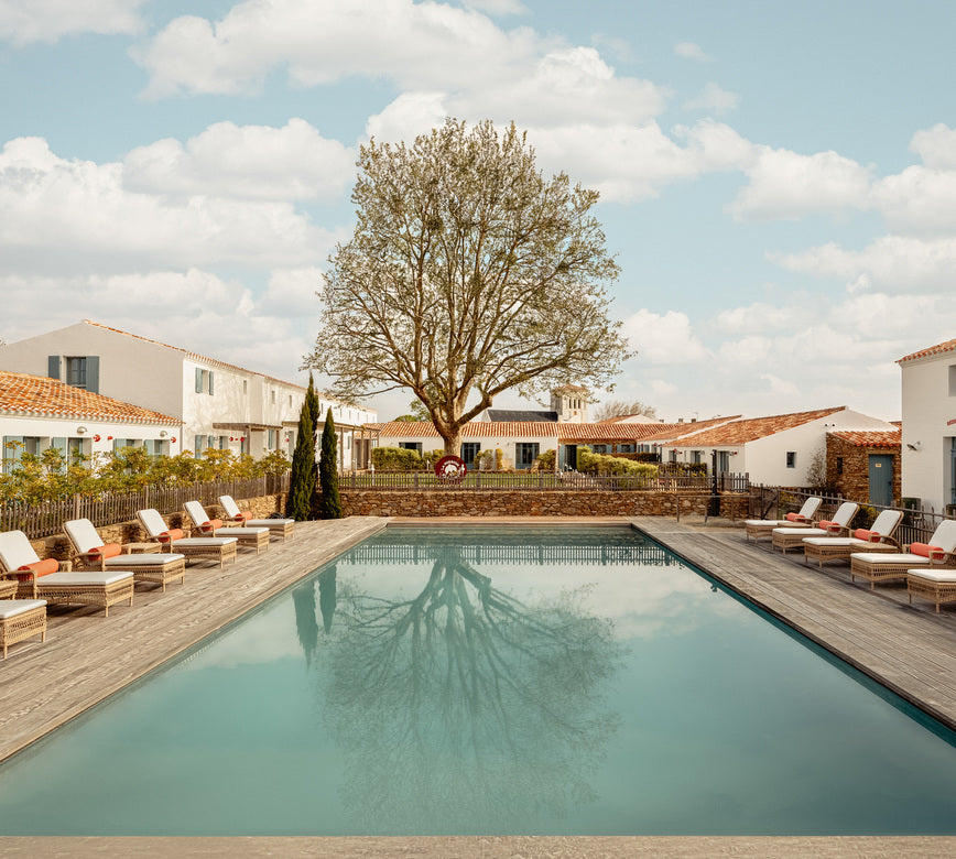 Bassin extérieur du spa NUXE à l’Hôtel La Mission, bordé d’une terrasse en bois aménagée de transats et parasols, dominé par un arbre central imposant se reflétant dans l’eau, entouré d’architectures traditionnelles aux toits en tuiles, sous une lumière naturelle apaisante offrant un cadre harmonieux, élégant et propice à la détente. 