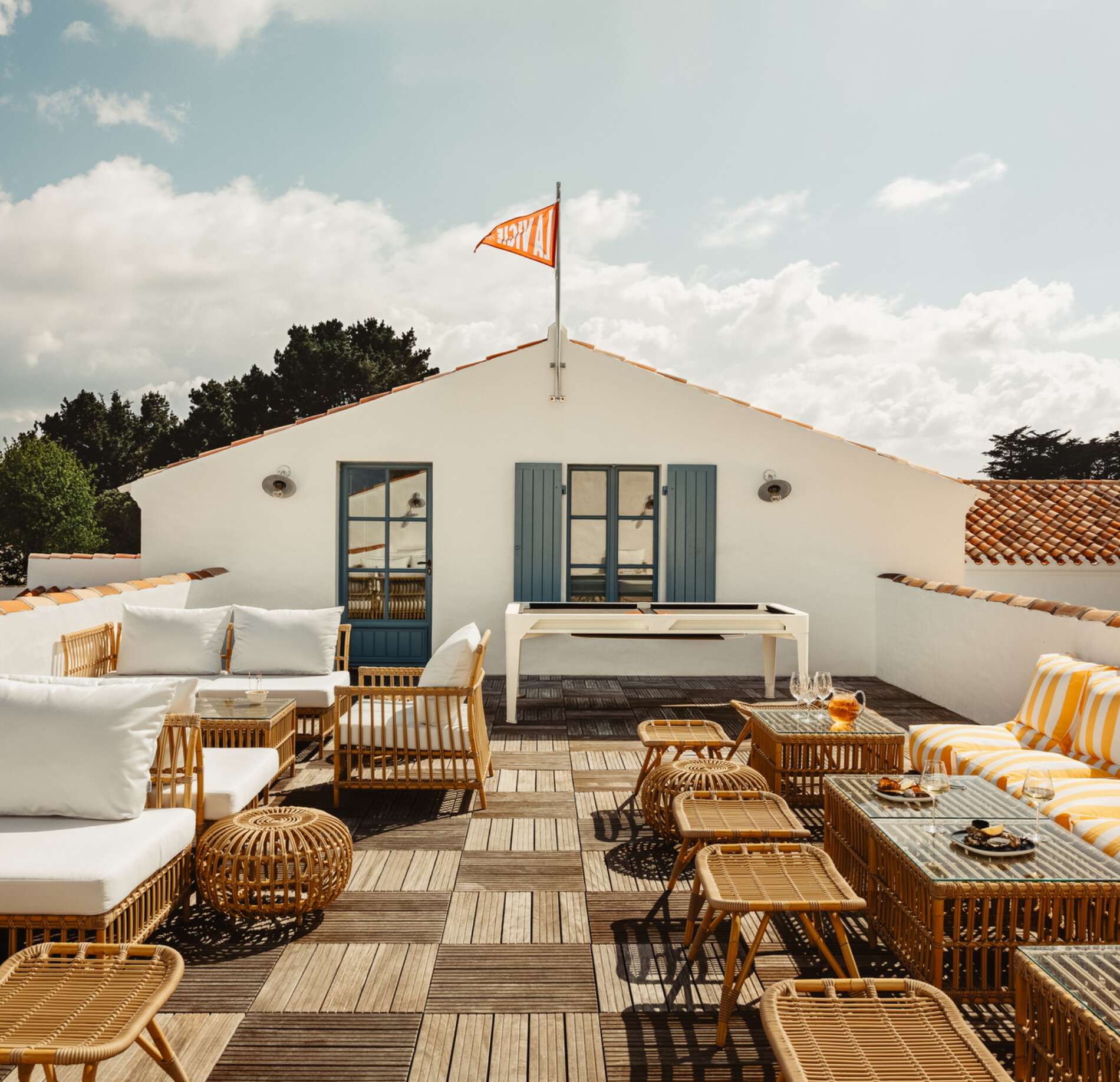 Terrasse extérieure élégante du spa NUXE à l’Hôtel La Mission, avec mobilier en fibres naturelles, canapés aux coussins clairs et touches orangées, table basse conviviale et façade blanche aux volets bleus sous un ciel lumineux, créant une atmosphère chaleureuse, raffinée et ensoleillée.
