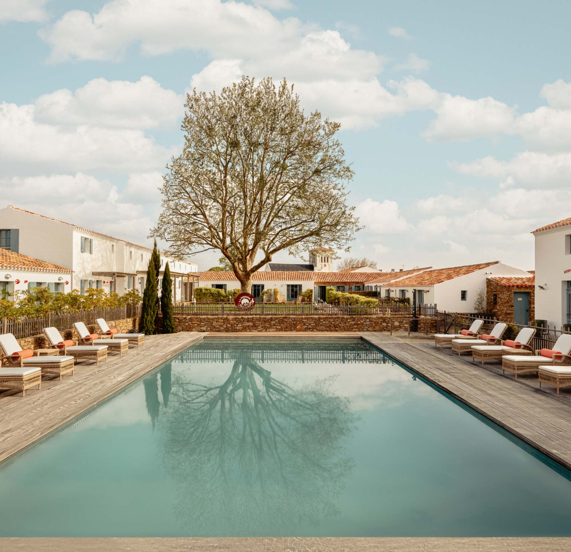 Piscine extérieure élégante du spa NUXE à l’Hôtel La Mission, entourée de transats confortables sur terrasse en bois, avec un grand arbre majestueux au centre et des bâtiments aux façades blanches typiques sous un ciel doux parsemé de nuages, créant une atmosphère paisible et raffinée.