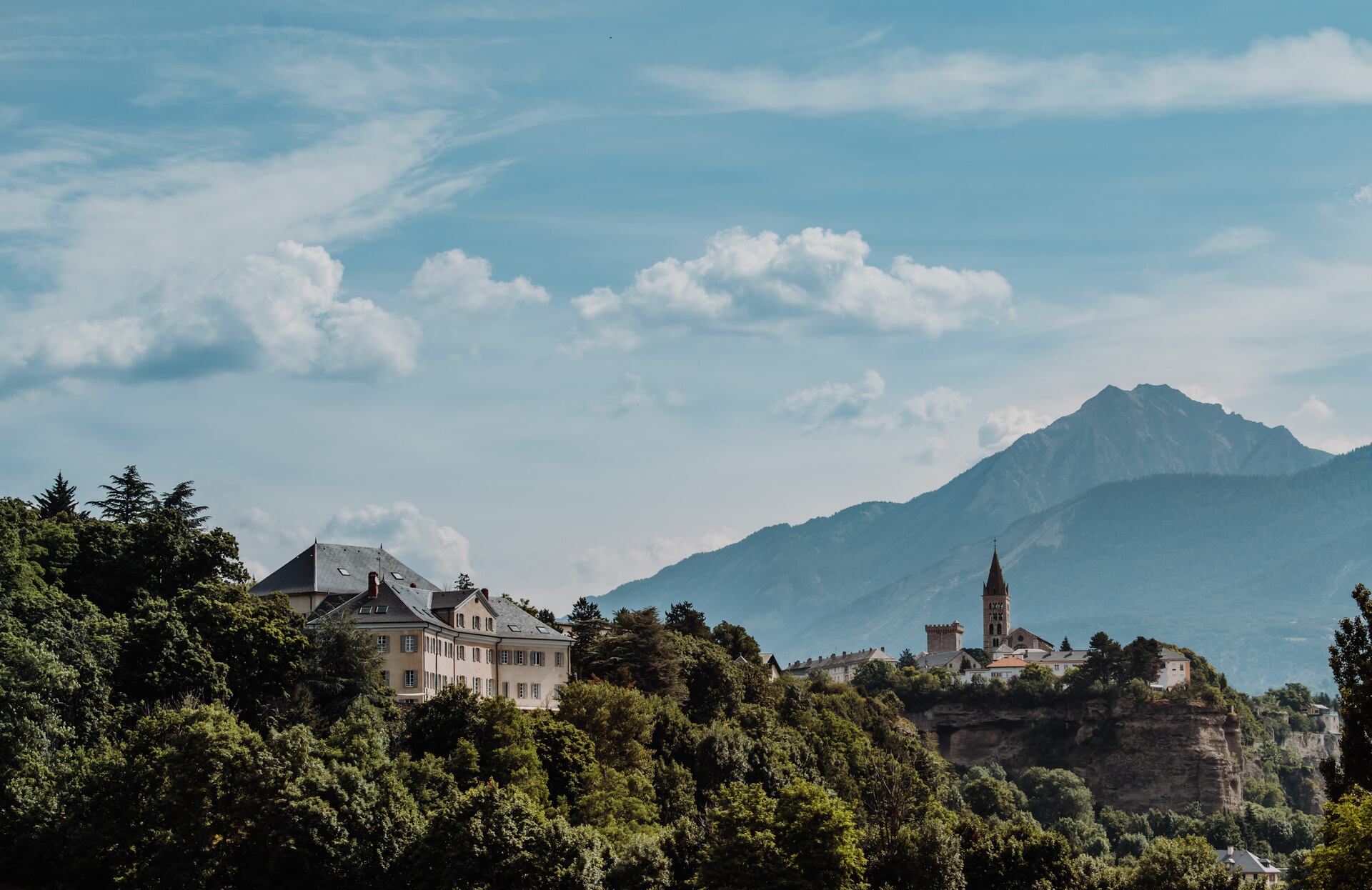 Une vue panoramique révèle un village perché sur une falaise, entouré d’une végétation dense, avec des bâtiments élégants dominés par un clocher finement élancé.
À l’arrière-plan, une chaîne de montagnes majestueuses se dessine sous un ciel lumineux, offrant un décor spectaculaire propice à une escapade mêlant nature et raffinement.