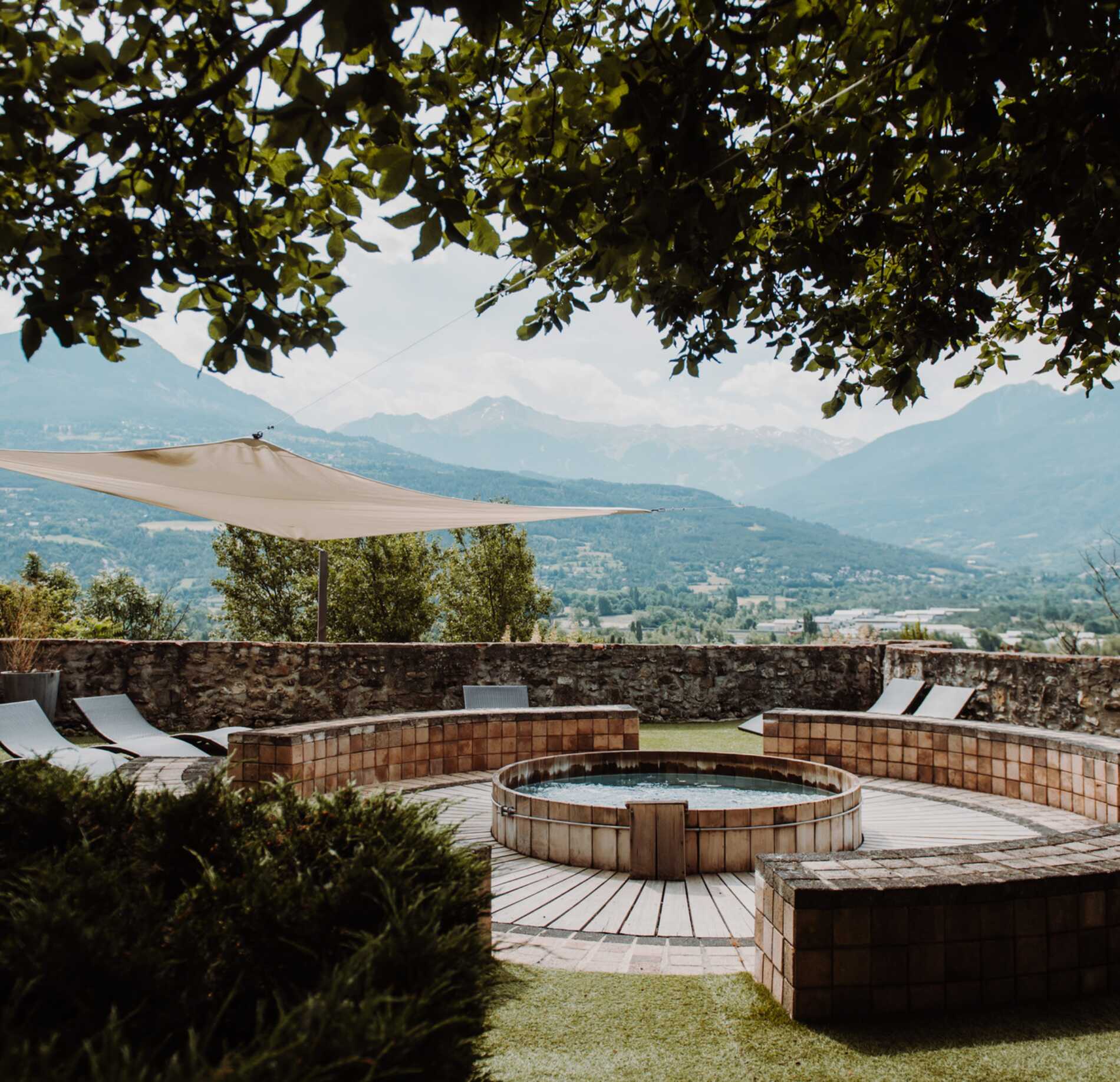Espace bien-être extérieur du spa NUXE à l’Hôtel La Robeyère, avec bain circulaire en bois encastré dans une terrasse surélevée, entouré de transats élégants et d’un muret en pierre, offrant une vue panoramique apaisante sur une vallée et des montagnes sous une lumière douce. 